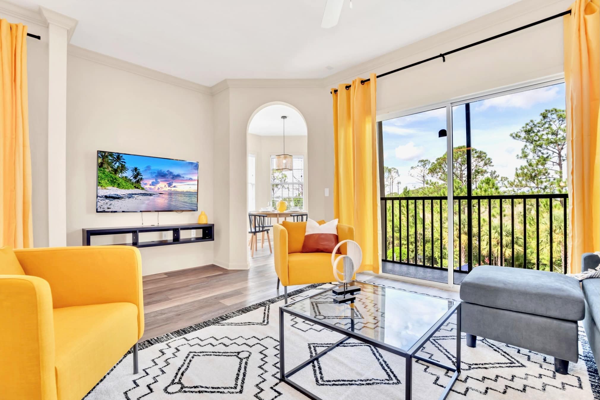 Living room with a wall-mounted TV, a yellow armchair, and a grey ottoman. A glass coffee table rests on a geometric rug, and a large sliding glass door opens to a balcony overlooking lush greenery.
