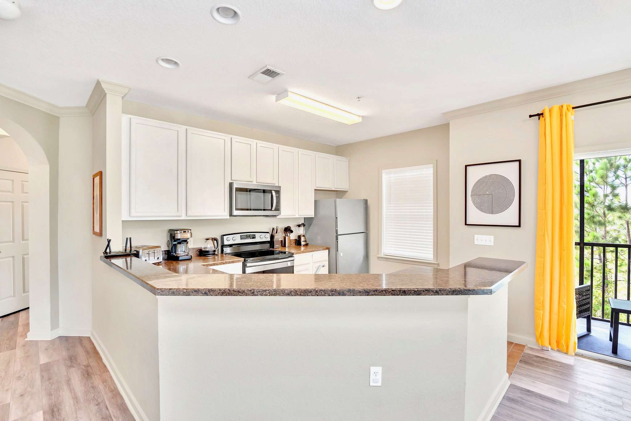 Modern kitchen with white shaker cabinets, stainless steel appliances, and brown granite countertops. A breakfast bar with seating separates the kitchen from the living area, offering a view towards the balcony.