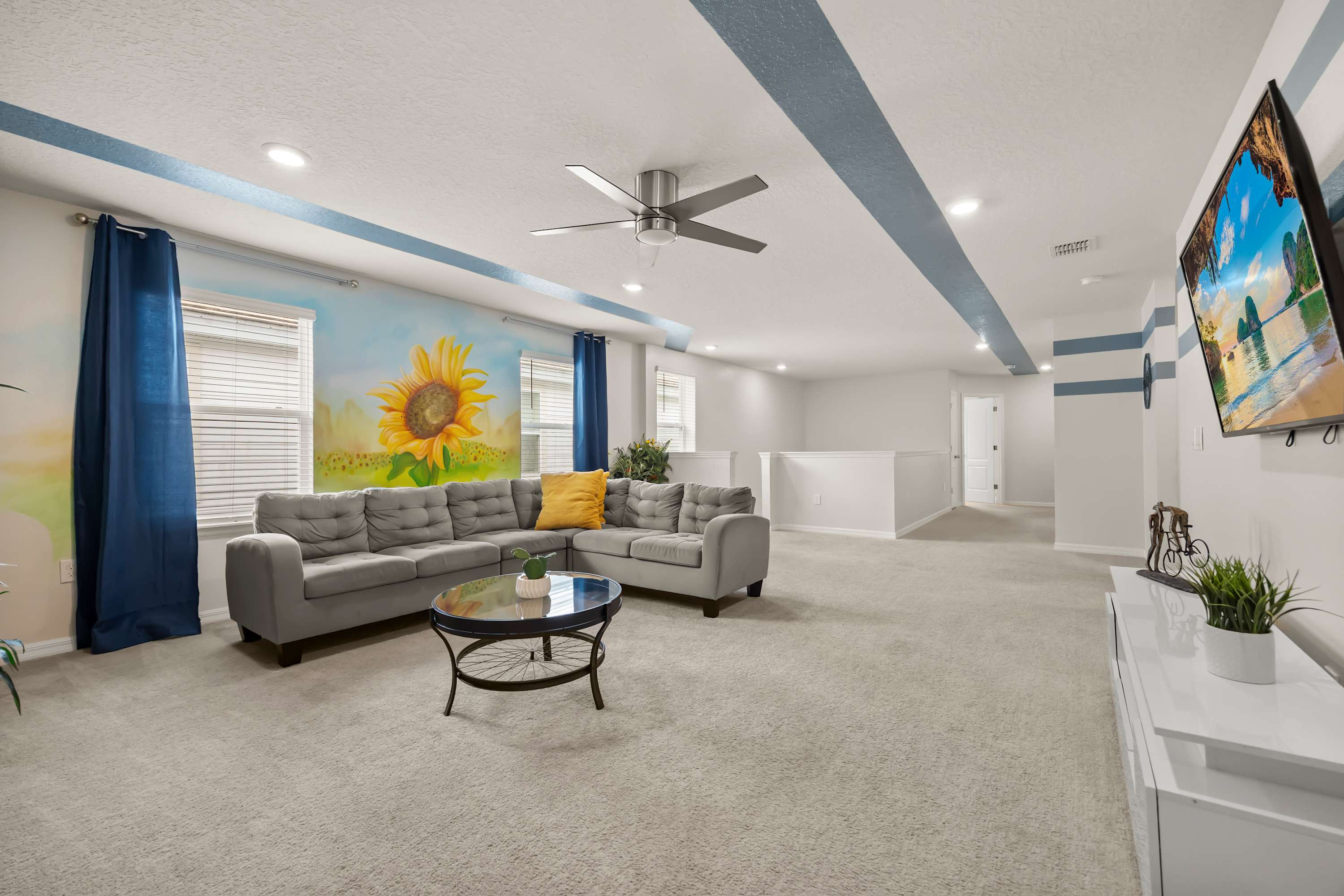 Expansive living room with a grey sectional sofa, a round glass coffee table, and a vibrant sunflower mural spanning the far wall, complemented by blue curtains and a ceiling fan.