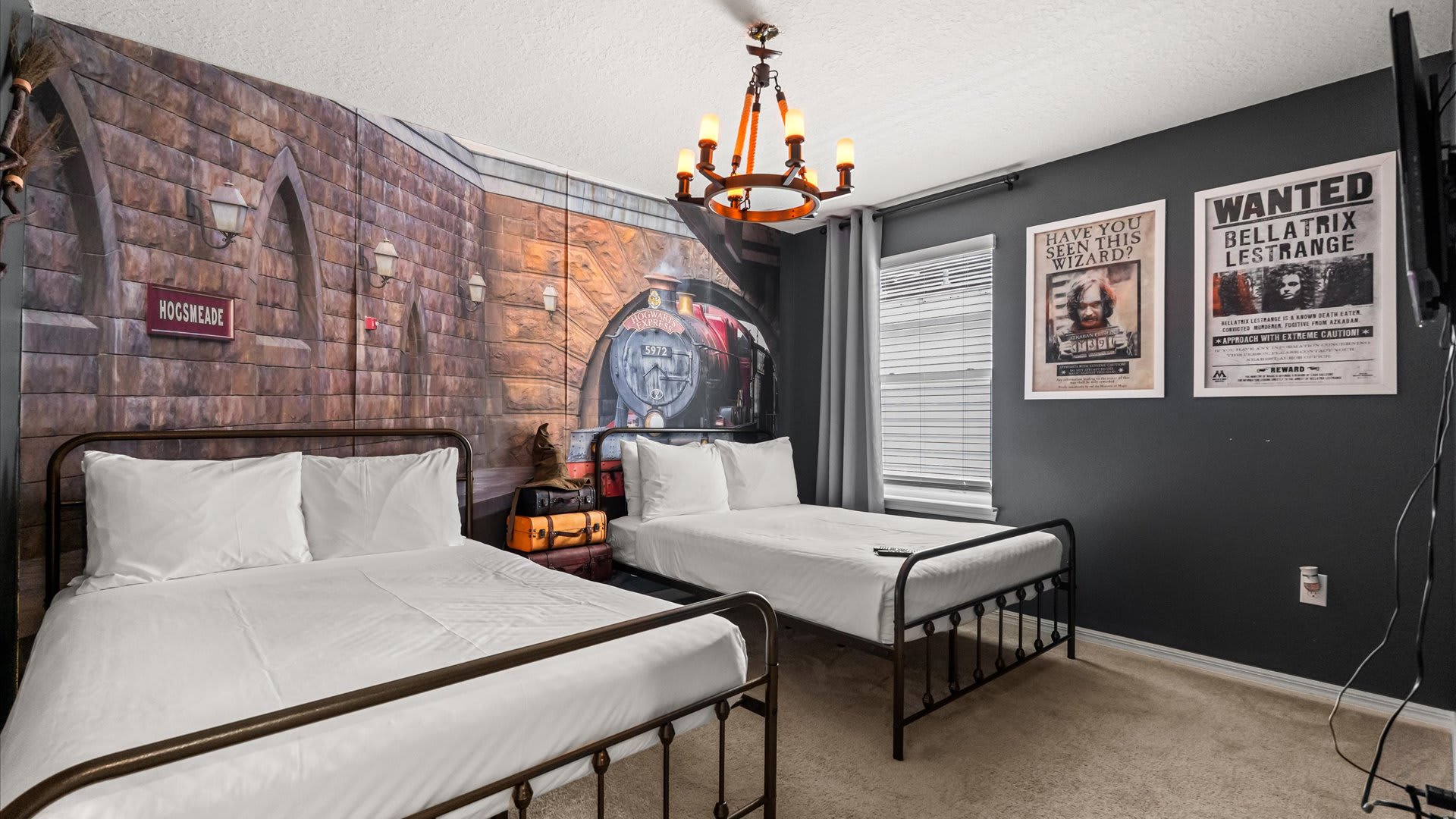 Themed bedroom with two single beds featuring dark metal frames and white bedding, set against a large mural depicting Hogsmeade station with the Hogwarts Express, under a rustic chandelier. Dark gray walls feature "Wanted" posters and a wall-mounted TV.