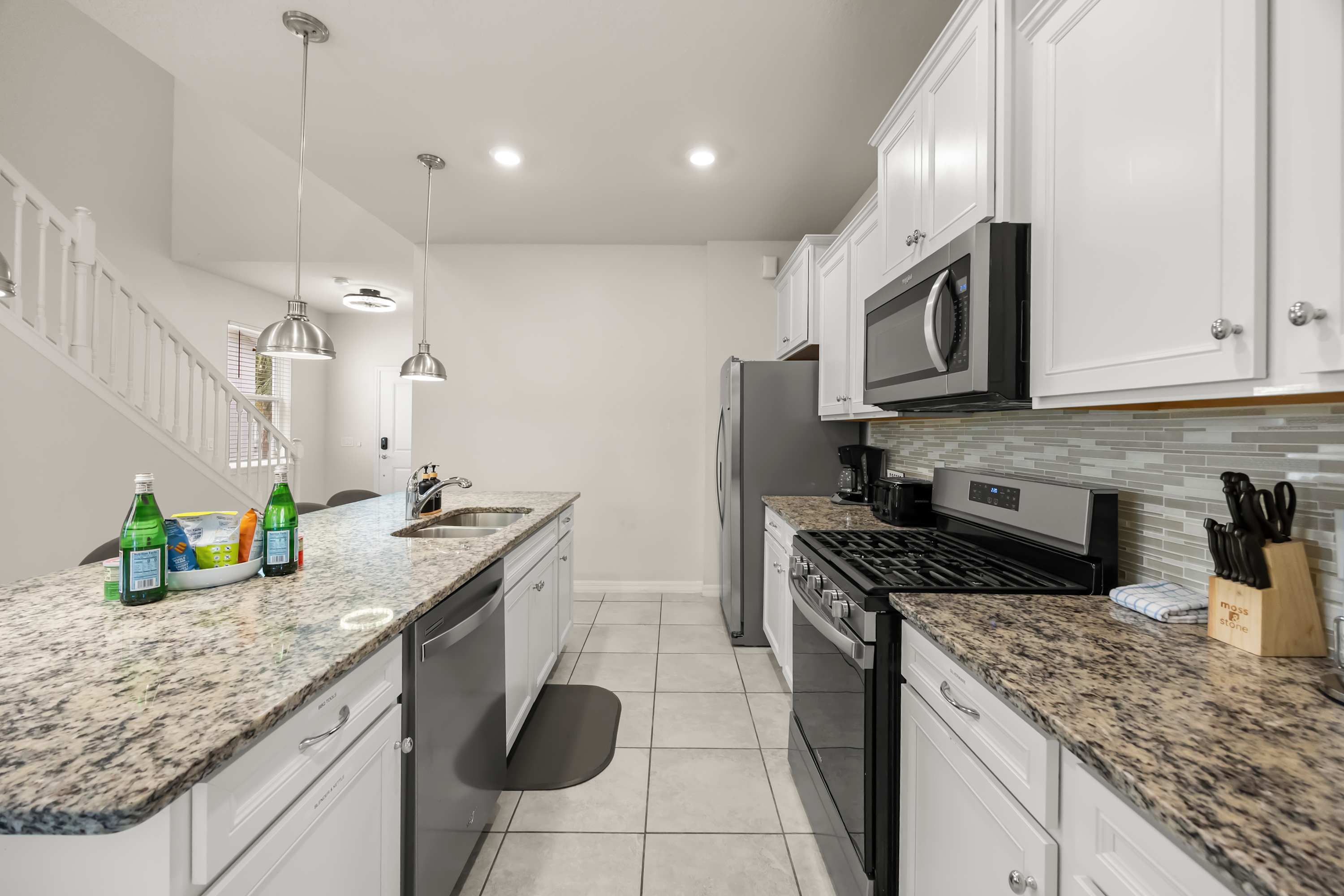 A galley-style kitchen with white cabinets, granite countertops, and stainless steel appliances, including a gas range and refrigerator, leading to a staircase in the background.