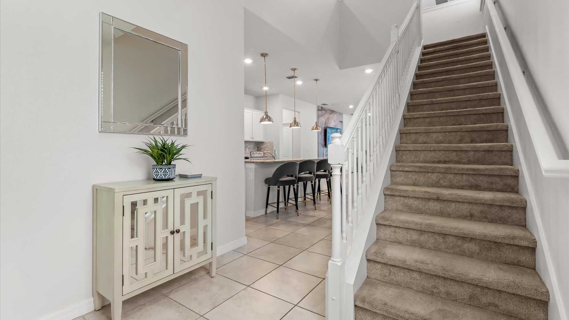 A bright foyer featuring a light-colored console table with a mirrored front and a potted plant, topped by a large rectangular mirror, with a carpeted staircase leading upstairs.