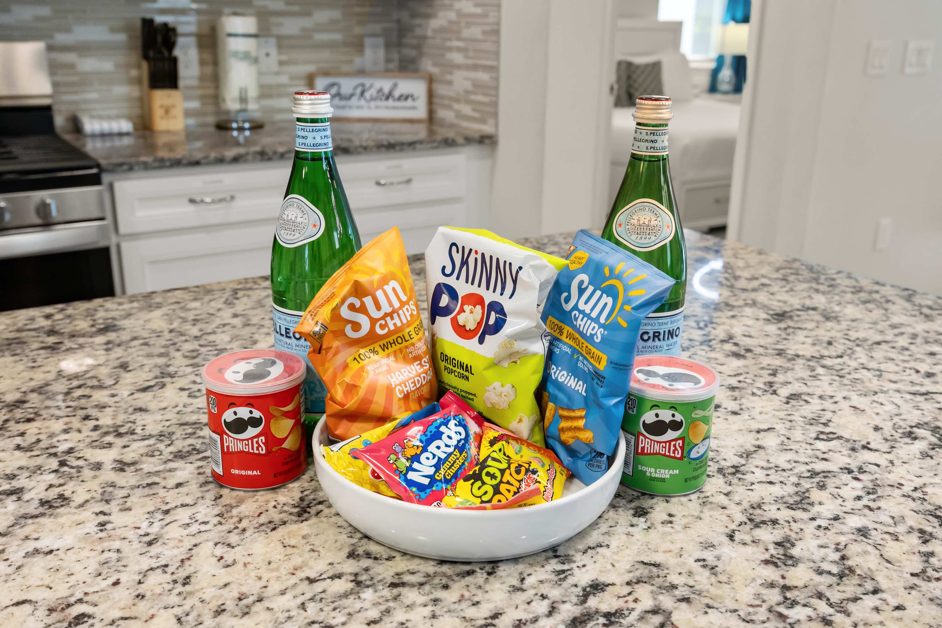 A close-up of a kitchen island with granite countertops, displaying a white bowl filled with various candies and chips, flanked by two bottles of San Pellegrino sparkling water and Pringles cans.
