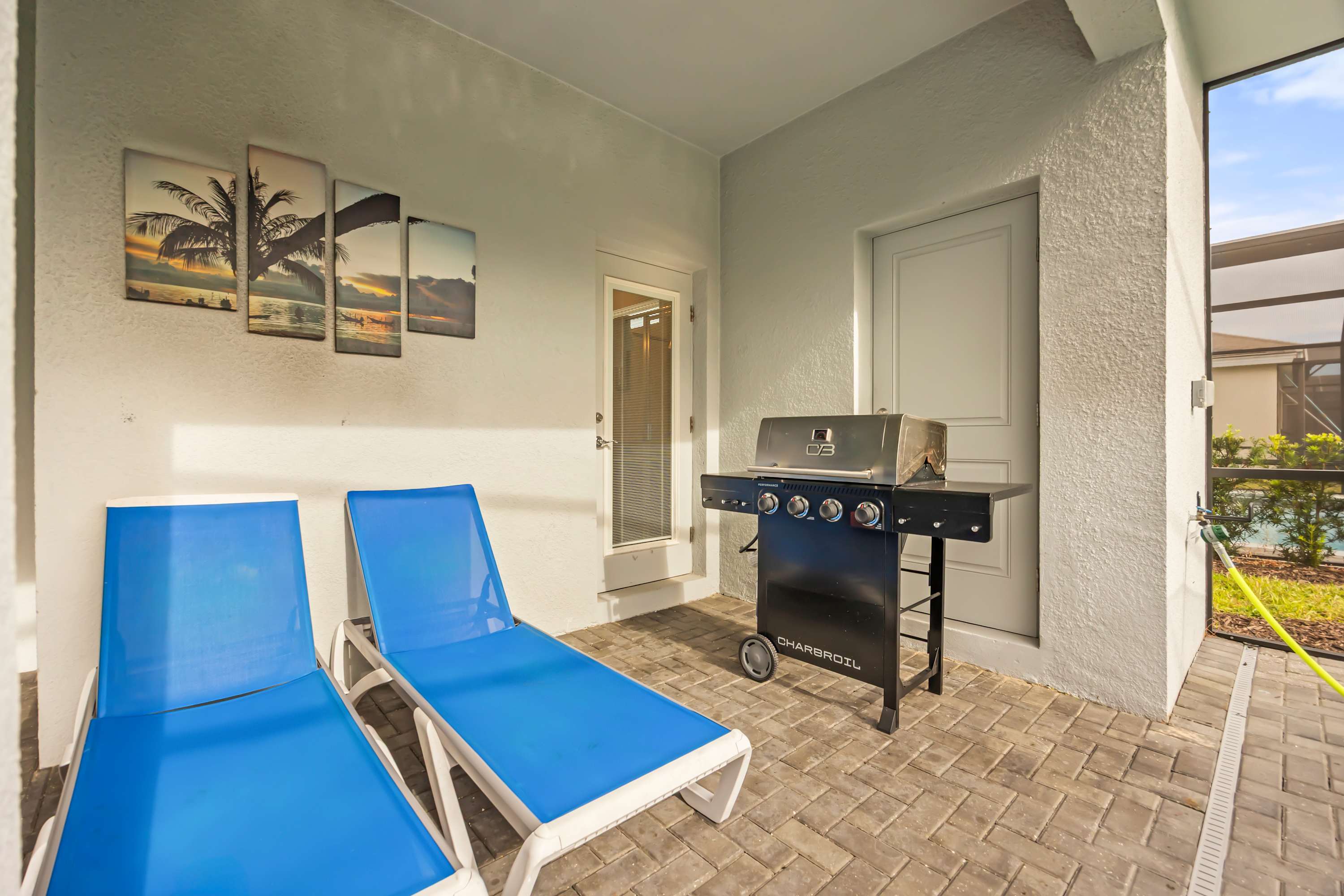Covered patio space featuring a stainless steel BBQ grill and two bright blue lounge chairs, with tropical-themed artwork adorning the wall.