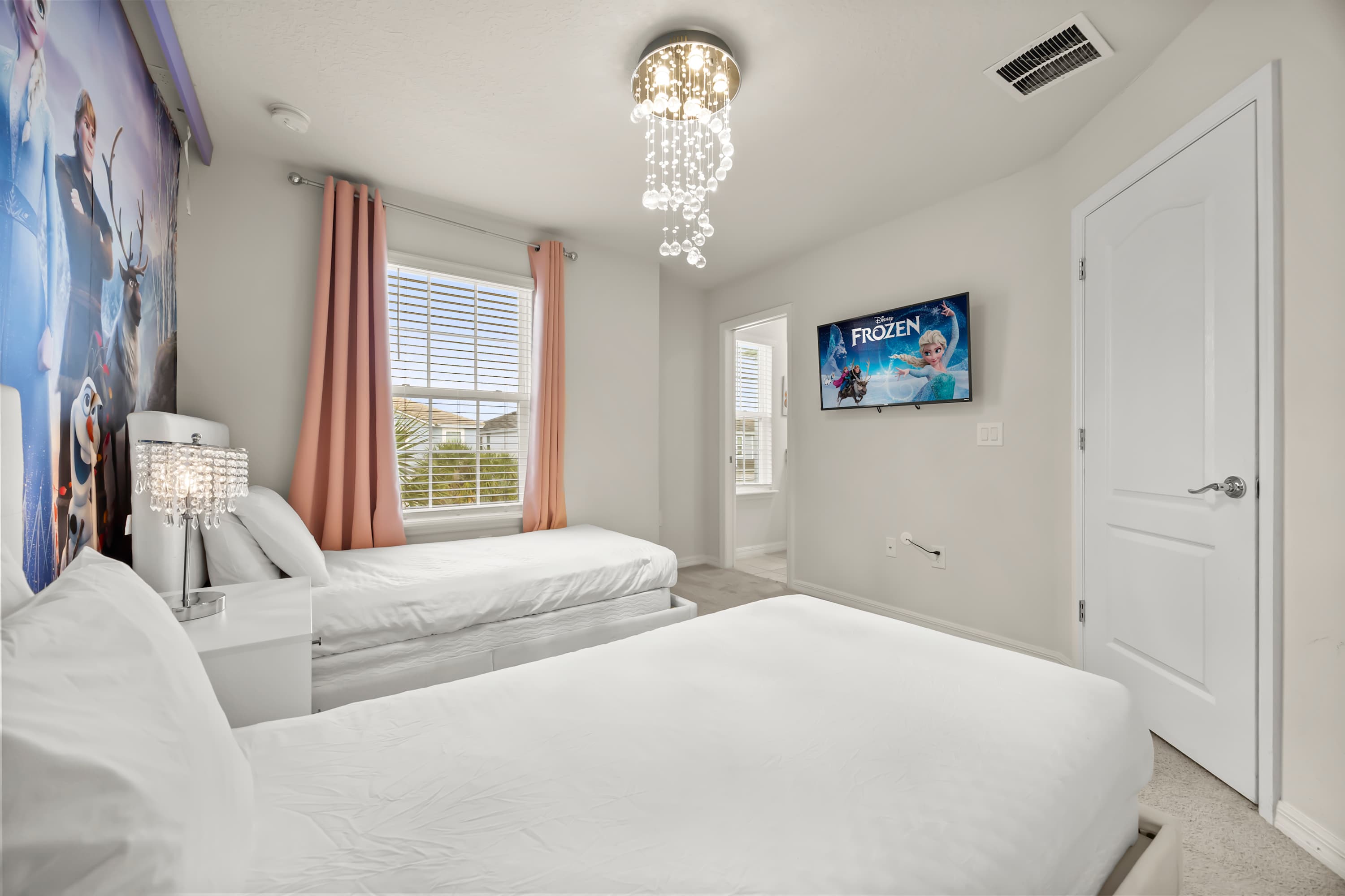A themed bedroom featuring two twin beds with white bedding, a "Frozen" mural on the left wall, and a wall-mounted TV displaying "Frozen." A modern crystal chandelier hangs from the ceiling, and a window with blinds is flanked by pink curtains.