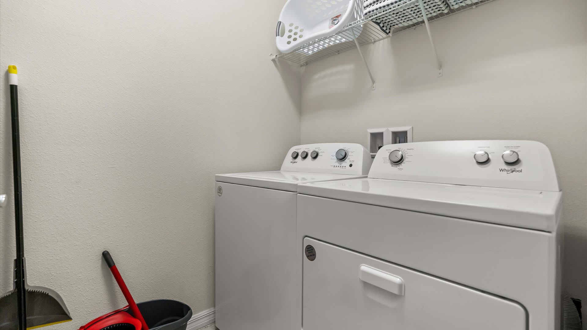 A functional laundry room featuring a white top-loading washing machine and a front-loading dryer. A wire shelf with a laundry basket is mounted above, and cleaning tools like a broom and dustpan are visible on the floor.