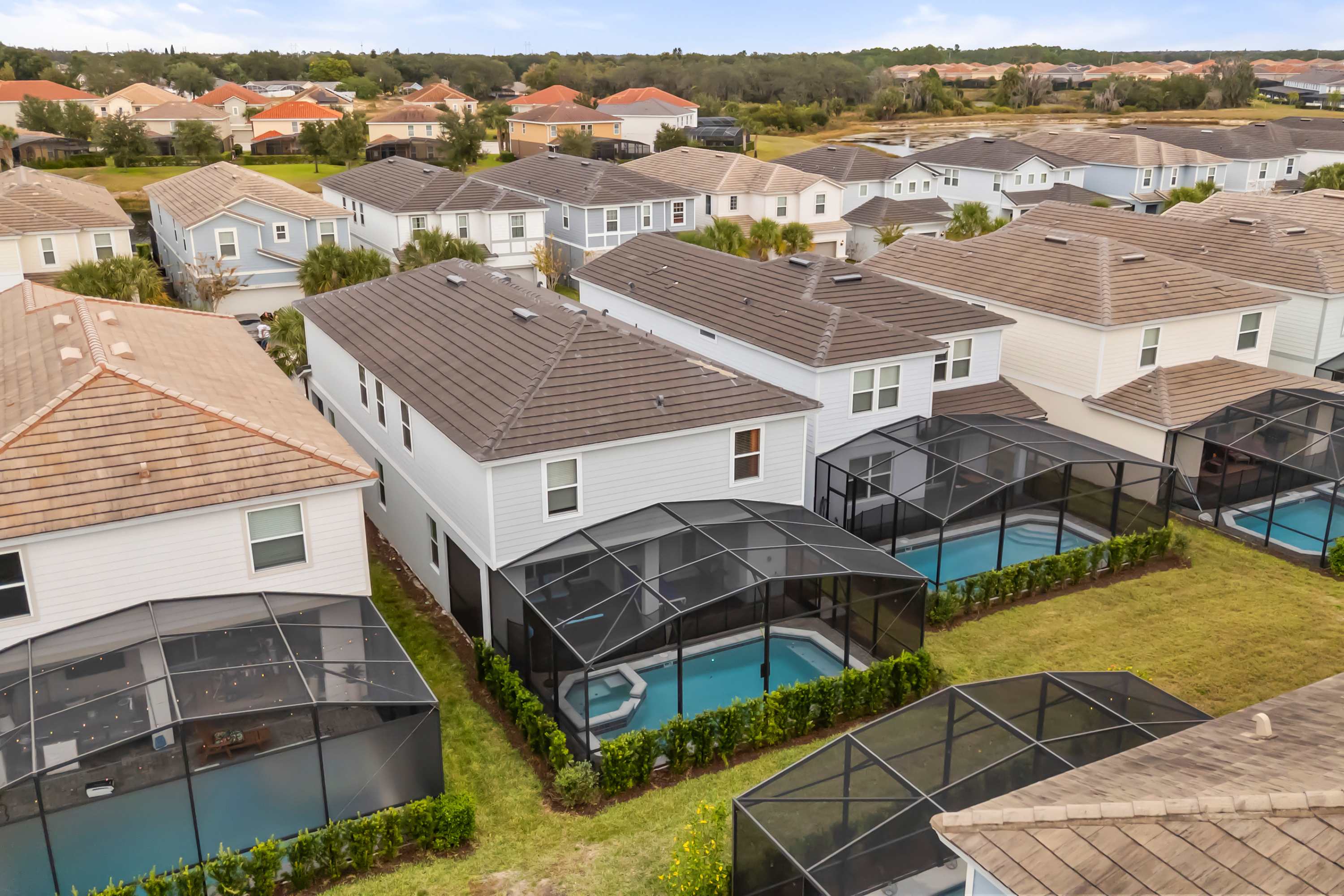 Aerial view of a residential neighborhood featuring several two-story houses with private screened-in pools in their backyards. The houses have light-colored siding and dark roofs, surrounded by green lawns and other similar properties.