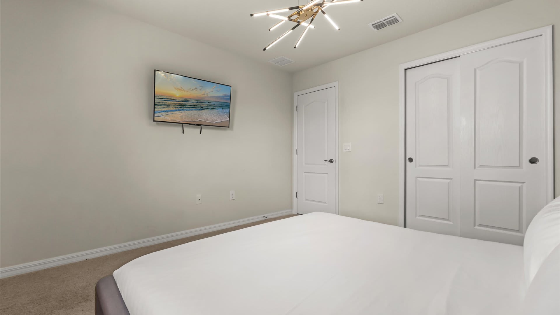 View of the bedroom showing a wall-mounted TV opposite the bed, a white closet with bi-fold doors, and a light-colored carpet, maintaining a clean and contemporary feel.