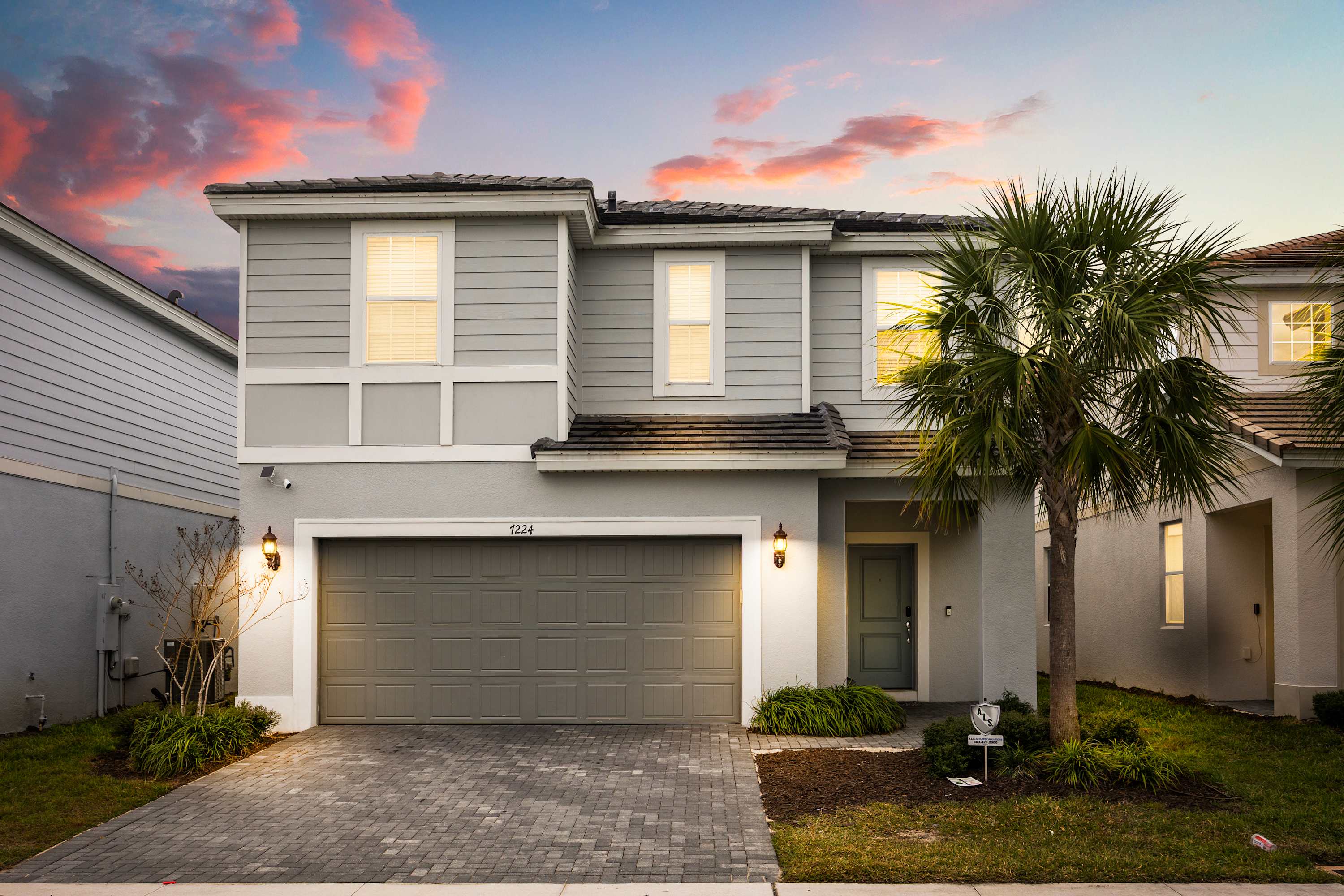 The front exterior of a modern two-story house with light gray siding and a dark gray garage door. A paved driveway leads to the garage, and a palm tree stands to the right of the entrance, all under a vibrant sunset sky.