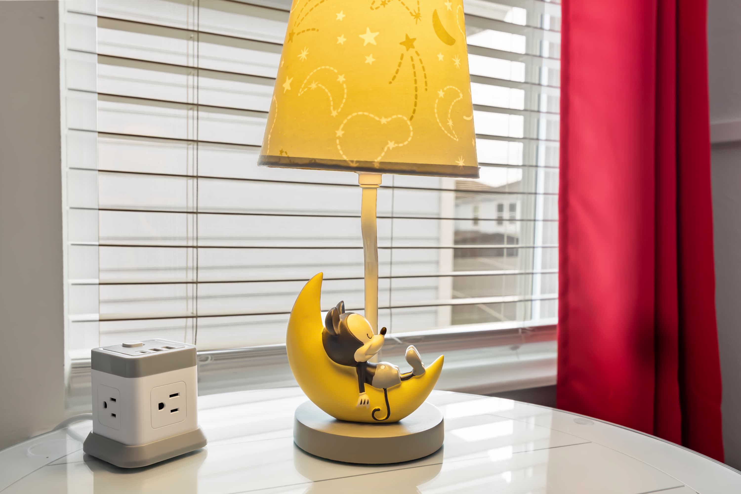 A close-up of a whimsical bedside table featuring a yellow moon and character lamp, a white power strip with USB ports, and a window with white blinds and red curtains.