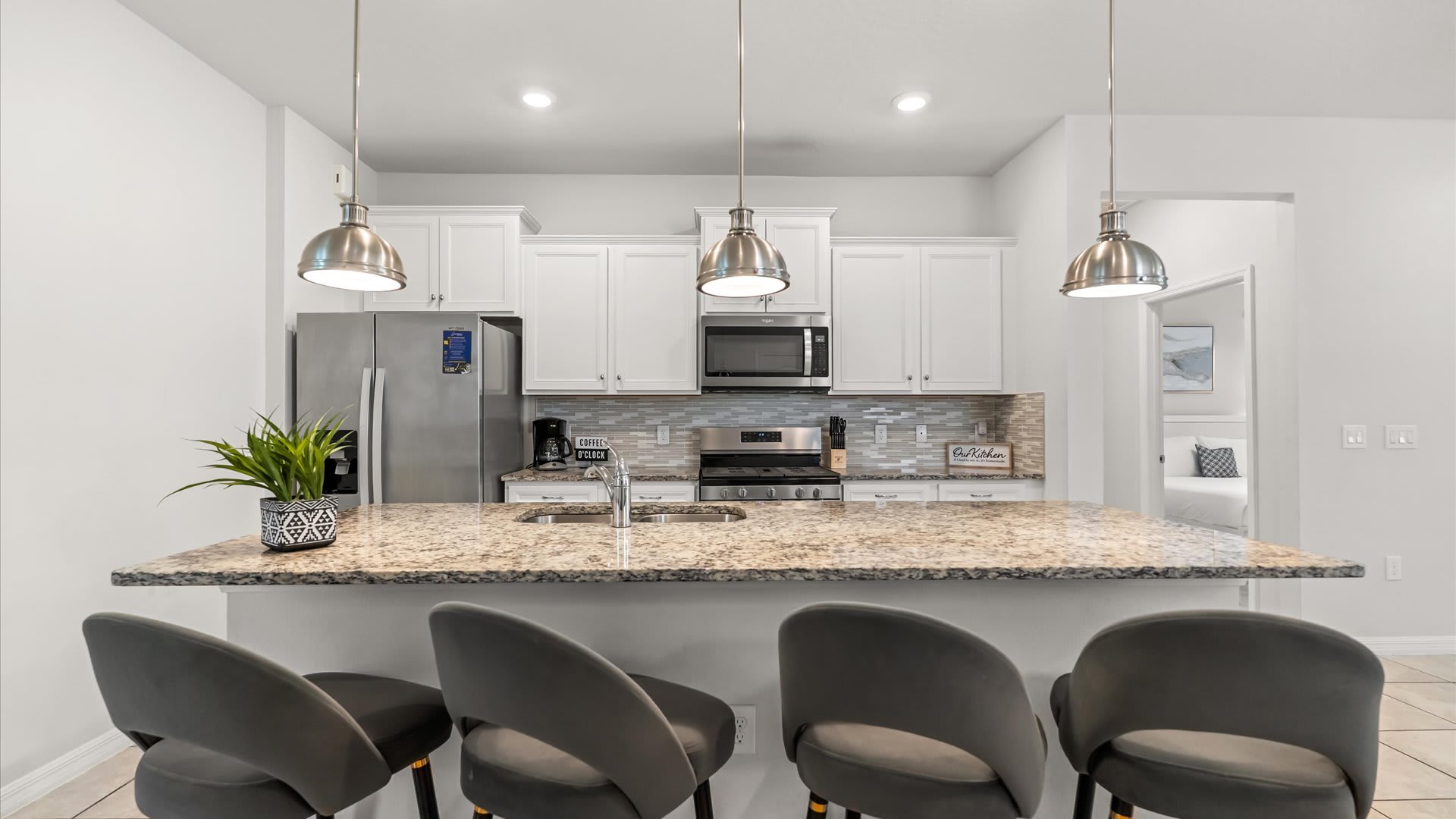 A bright kitchen featuring white cabinets, granite countertops, stainless steel appliances, and a large island with a sink and four gray bar stools, lit by three brushed nickel pendant lights.