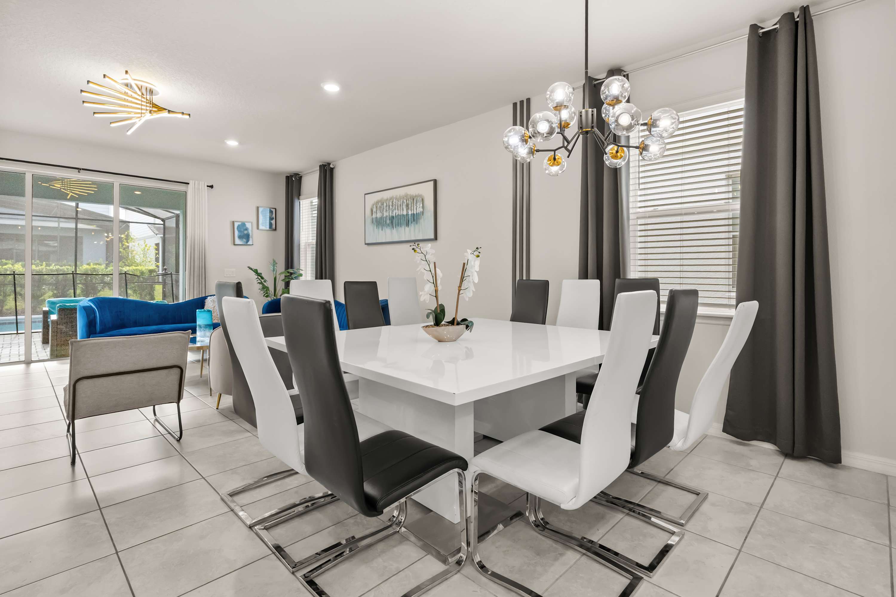 Modern dining area featuring a large white square dining table with seating for eight, comprised of black and white chairs, under a contemporary chandelier, adjacent to a living area.