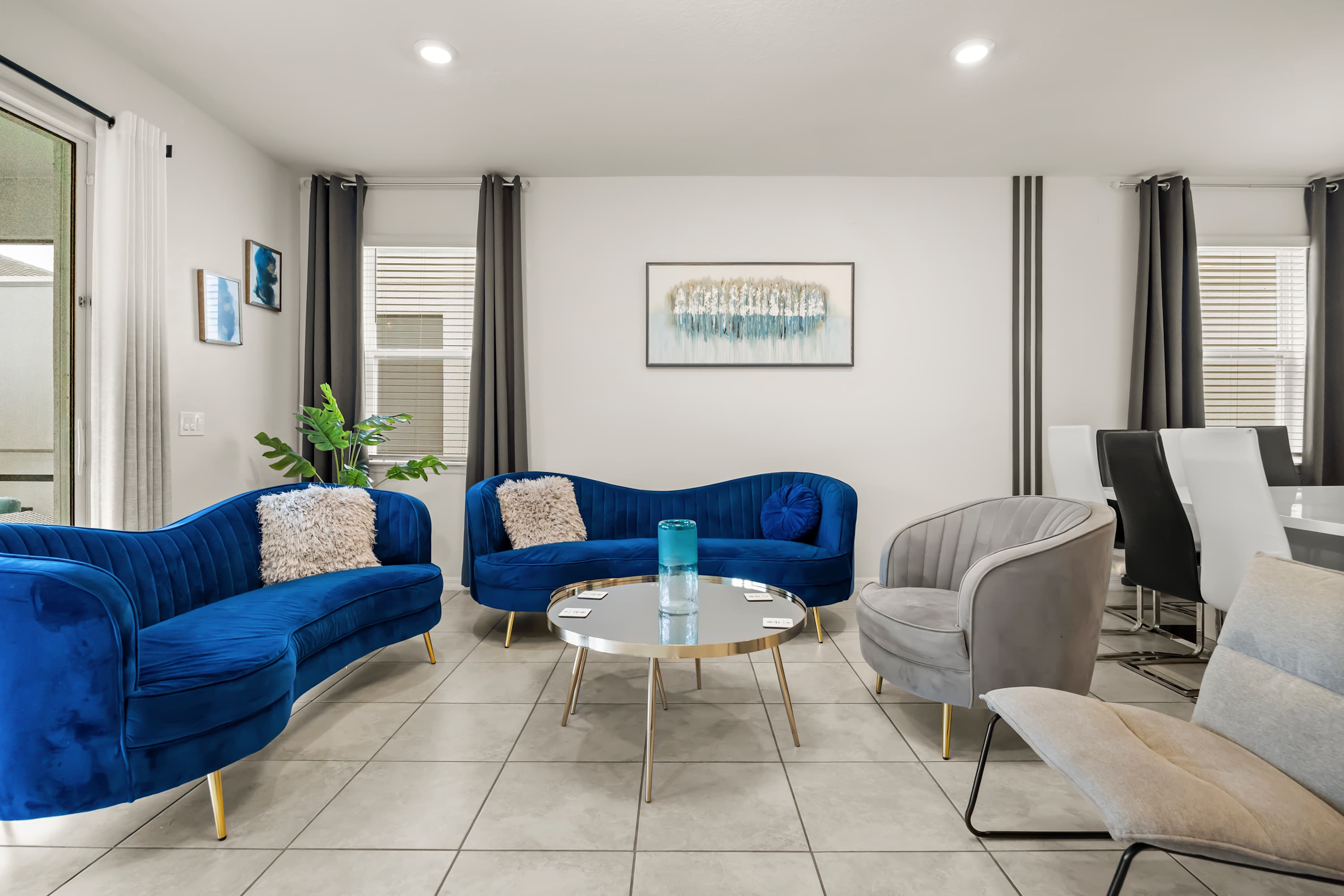 Elegant living room with light tiled flooring, two curved blue velvet sofas with gold legs, a round glass coffee table, and large windows with dark curtains.