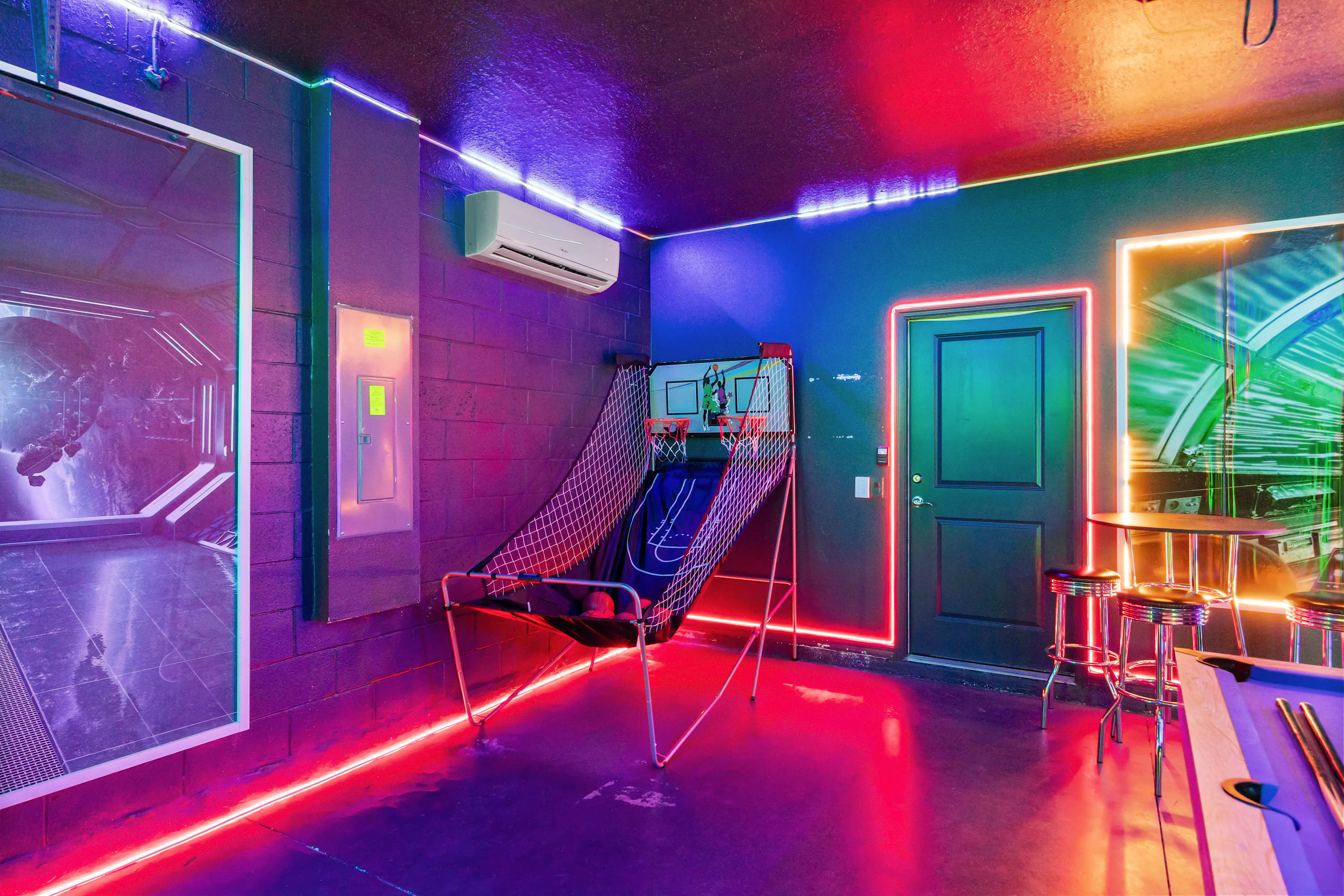 Vibrant game room with dark walls illuminated by red and blue neon lights, featuring an arcade basketball game, a pool table, and a high-top table with stools.