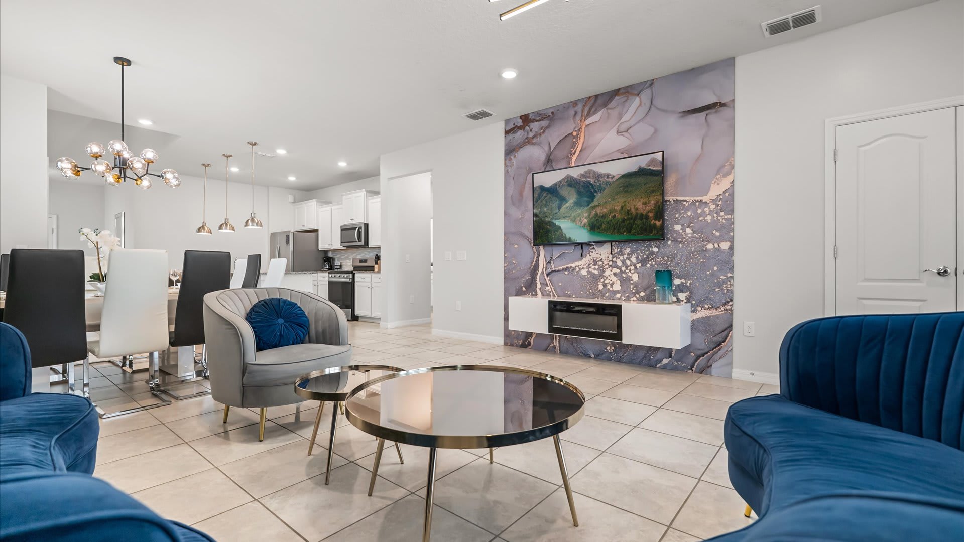 Modern living room featuring a large flat-screen TV mounted on a striking marble-patterned accent wall with a built-in electric fireplace, flanked by blue velvet sofas and grey armchairs.