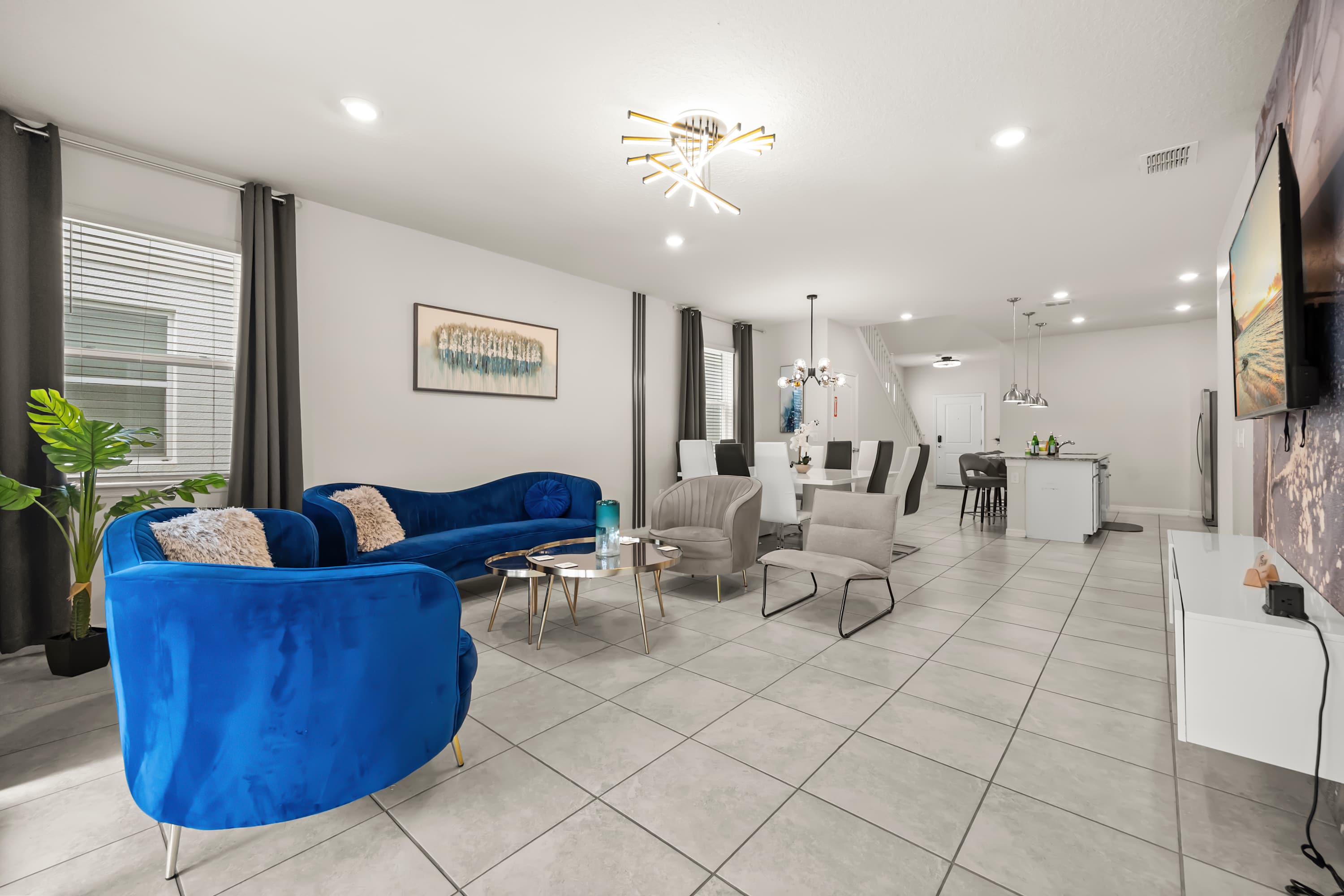 Spacious open-concept living area with light tiled floors, featuring two blue velvet sofas, a modern coffee table, and a view towards the dining area and kitchen.
