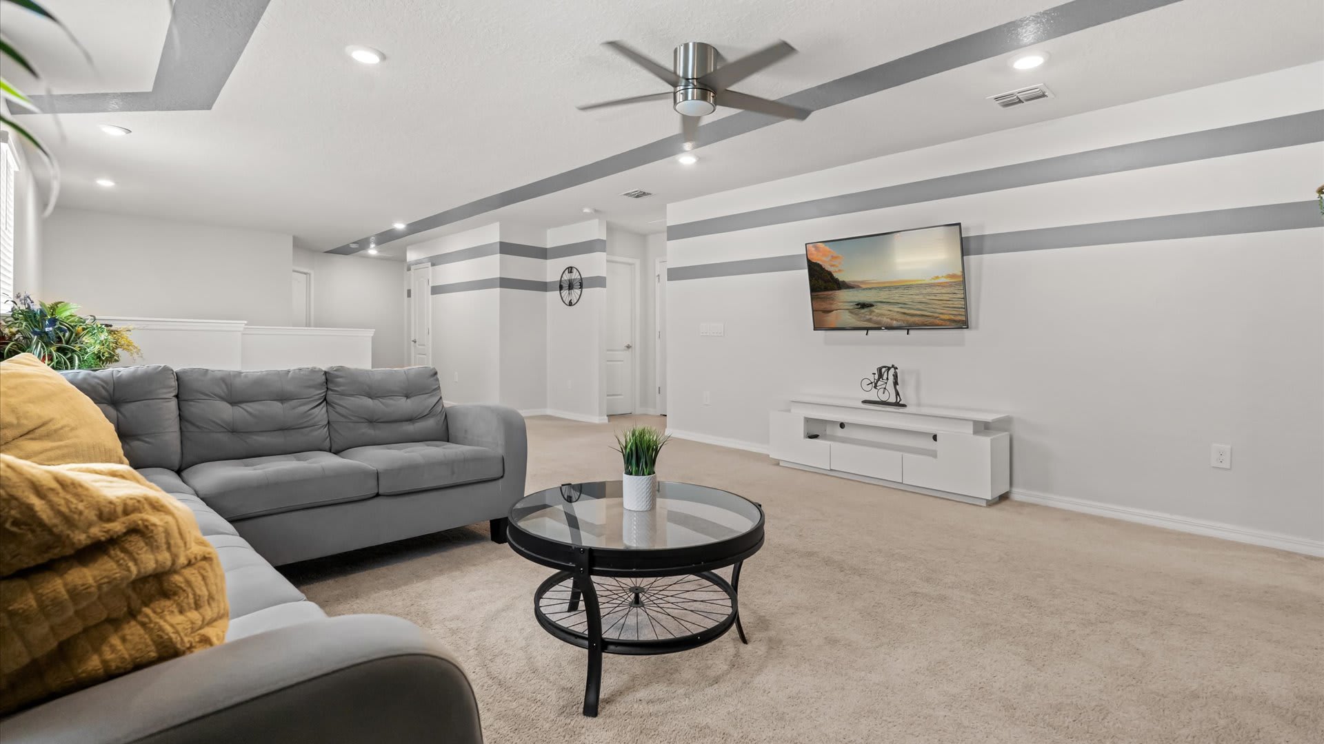 A modern living area with a large grey sectional sofa, a round glass coffee table, and a wall-mounted TV, featuring a white ceiling with bold grey stripes and recessed lighting.