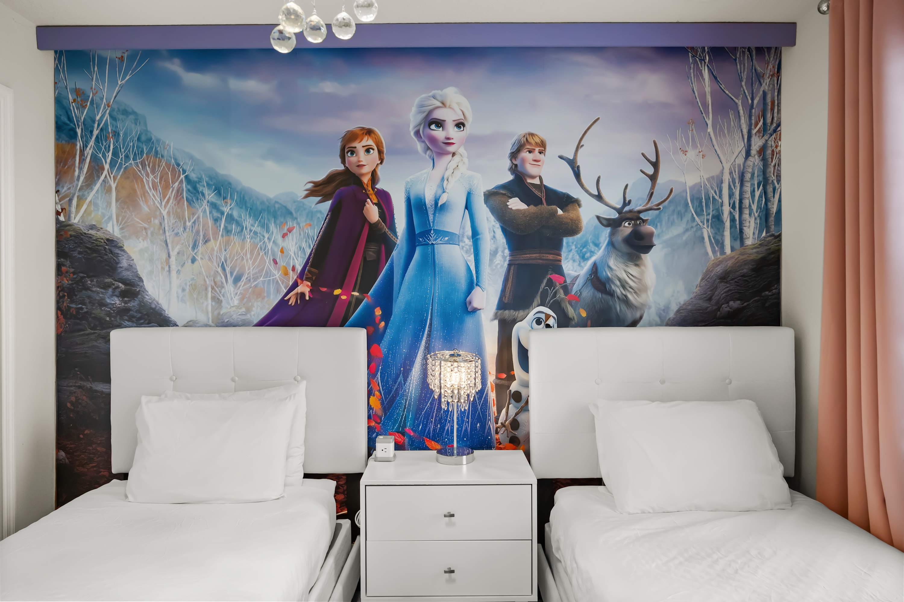 Frozen-themed bedroom featuring two white single beds with white bedding, a shared white nightstand with a decorative lamp, and a vibrant wall mural of Elsa, Anna, Kristoff, and Sven.