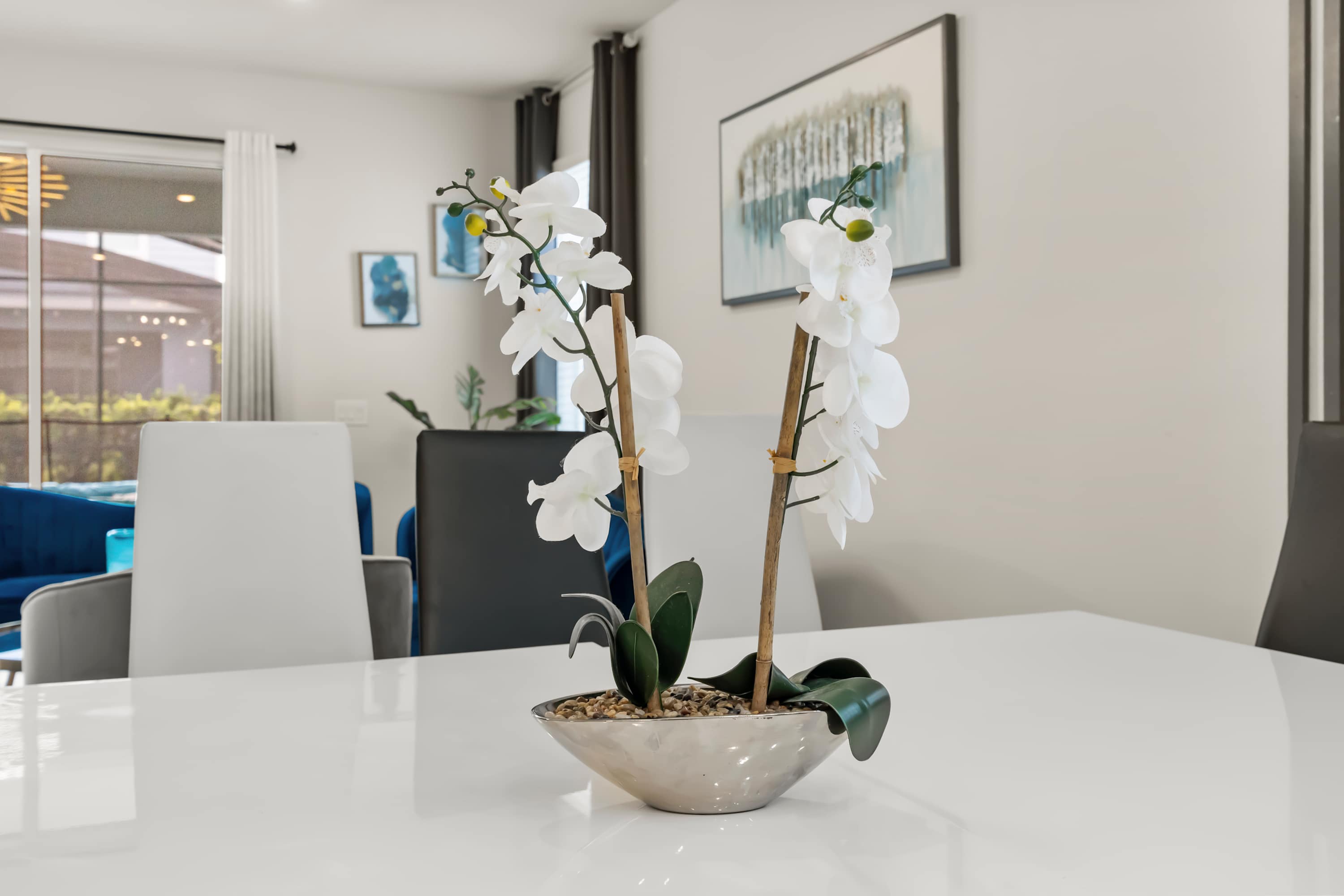 A close-up of a white dining table featuring a decorative centerpiece with two artificial white orchid plants in a silver bowl, set against a backdrop of modern dining chairs and a framed abstract artwork.