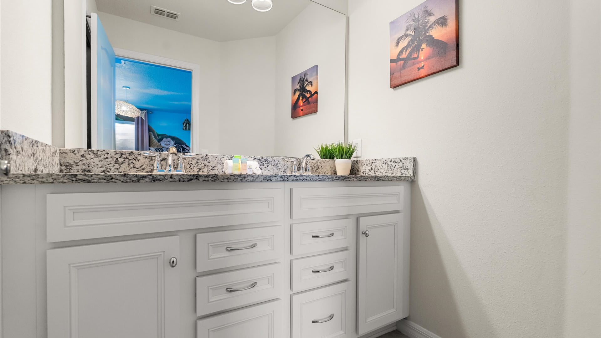 A bright en-suite bathroom featuring a double vanity with granite countertops, white cabinetry, and decorative tropical-themed wall art.