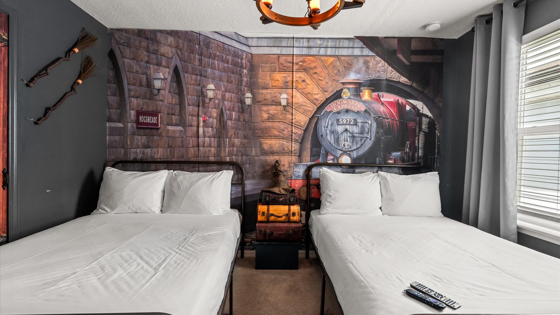 Themed bedroom with two single beds, each with dark metal frames and white bedding, positioned against a detailed mural of Hogsmeade station and the Hogwarts Express, with a vintage-style chandelier overhead.