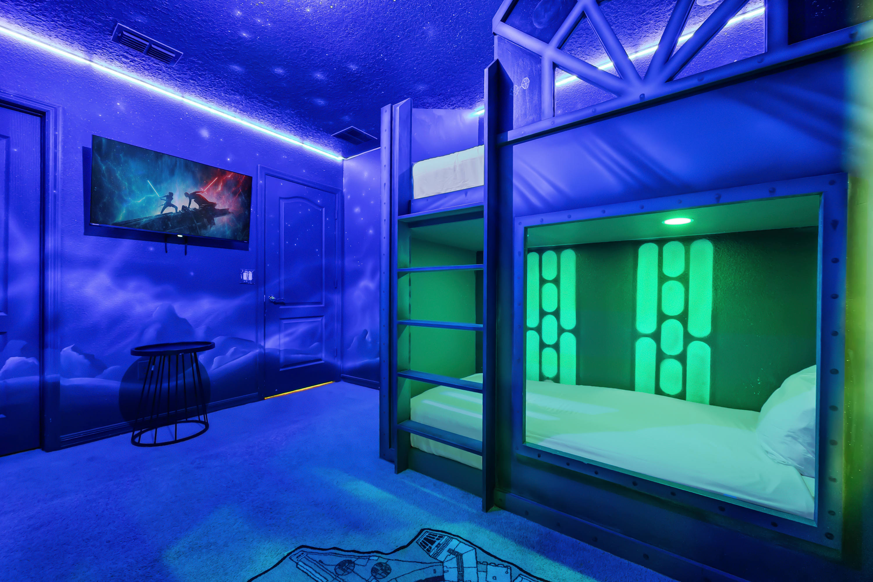 Star Wars themed bedroom with blue ambient lighting, featuring custom-built bunk beds with green illuminated interiors, a wall-mounted TV, and a Millennium Falcon rug on the carpeted floor.
