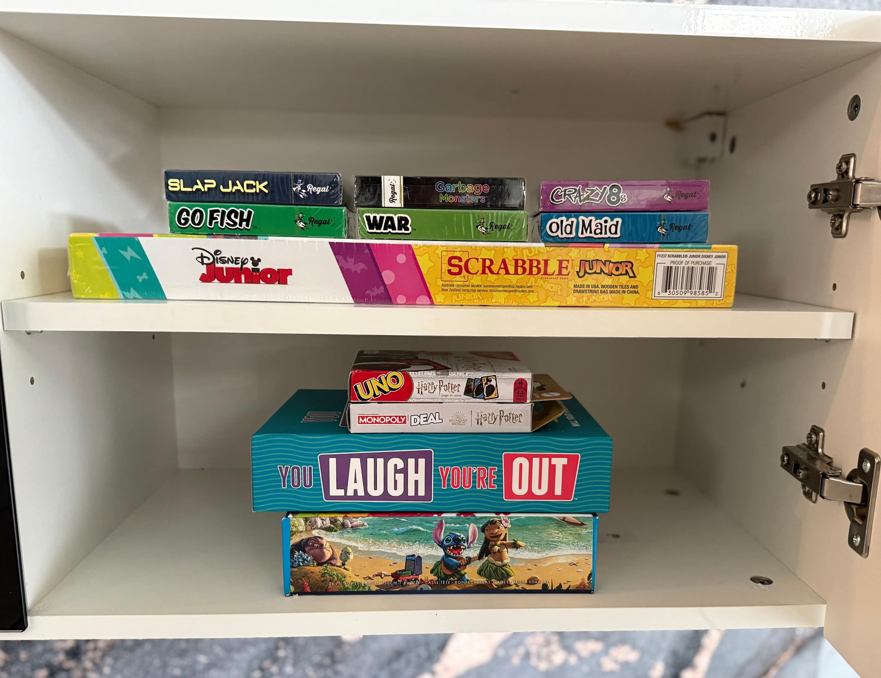 White cabinet shelves neatly stocked with a variety of board games and card games, including Scrabble Junior, UNO, Monopoly Deal, and classic card decks.