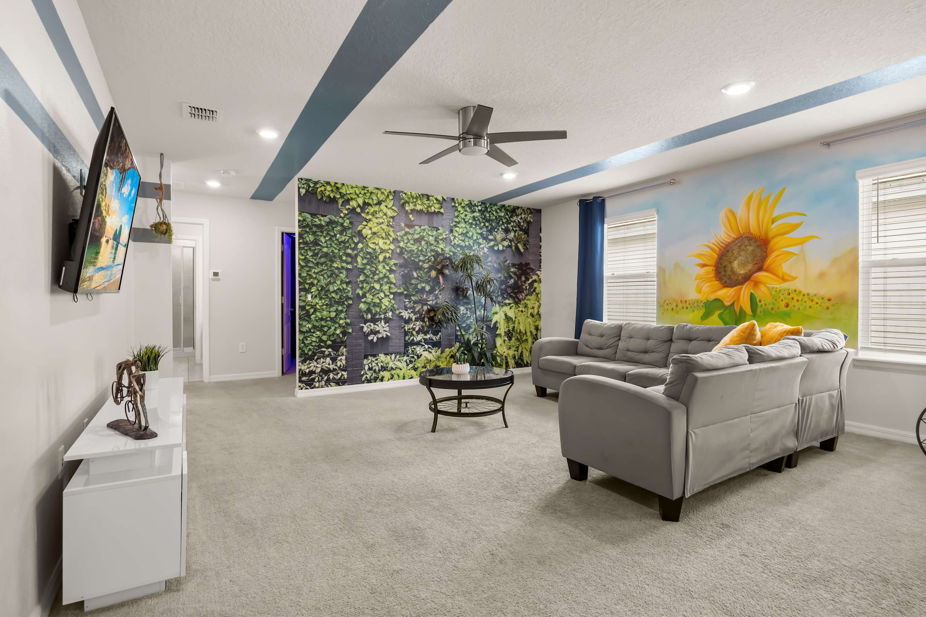 A spacious game room with light gray carpet, two large gray sofas, a round coffee table, a wall-mounted TV, and unique decor including a green plant wall and a vibrant sunflower mural.