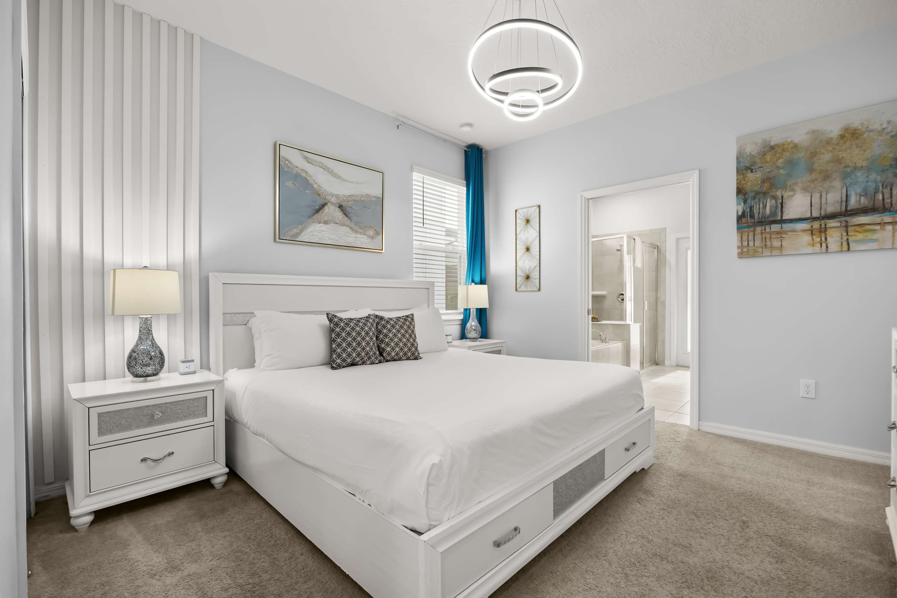 Bright bedroom featuring a white queen bed with storage drawers, two white nightstands with lamps, and a modern circular chandelier, with a door leading to an en-suite bathroom.
