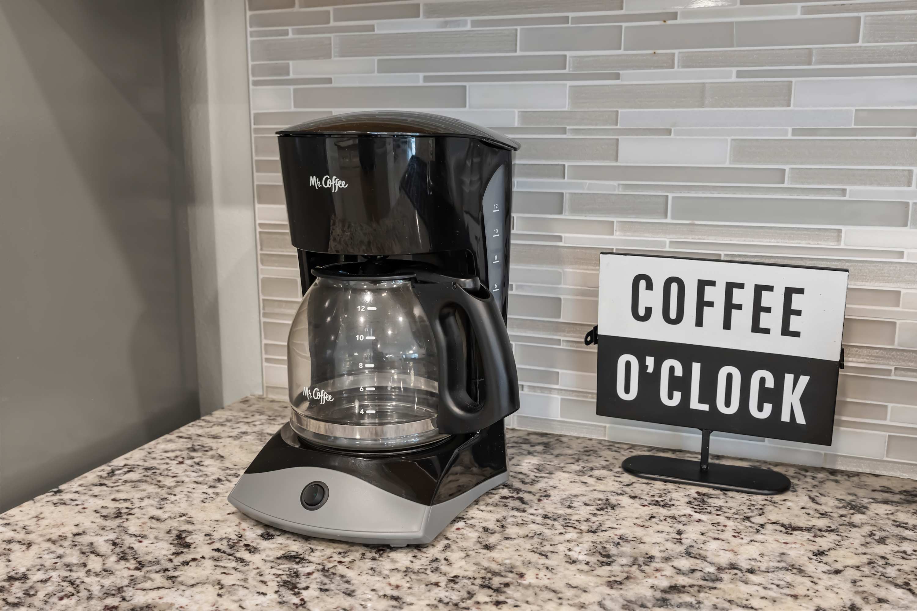A close-up of a black Mr. Coffee drip coffee maker on a granite countertop, next to a decorative sign that reads "COFFEE O'CLOCK" against a gray subway tile backsplash.