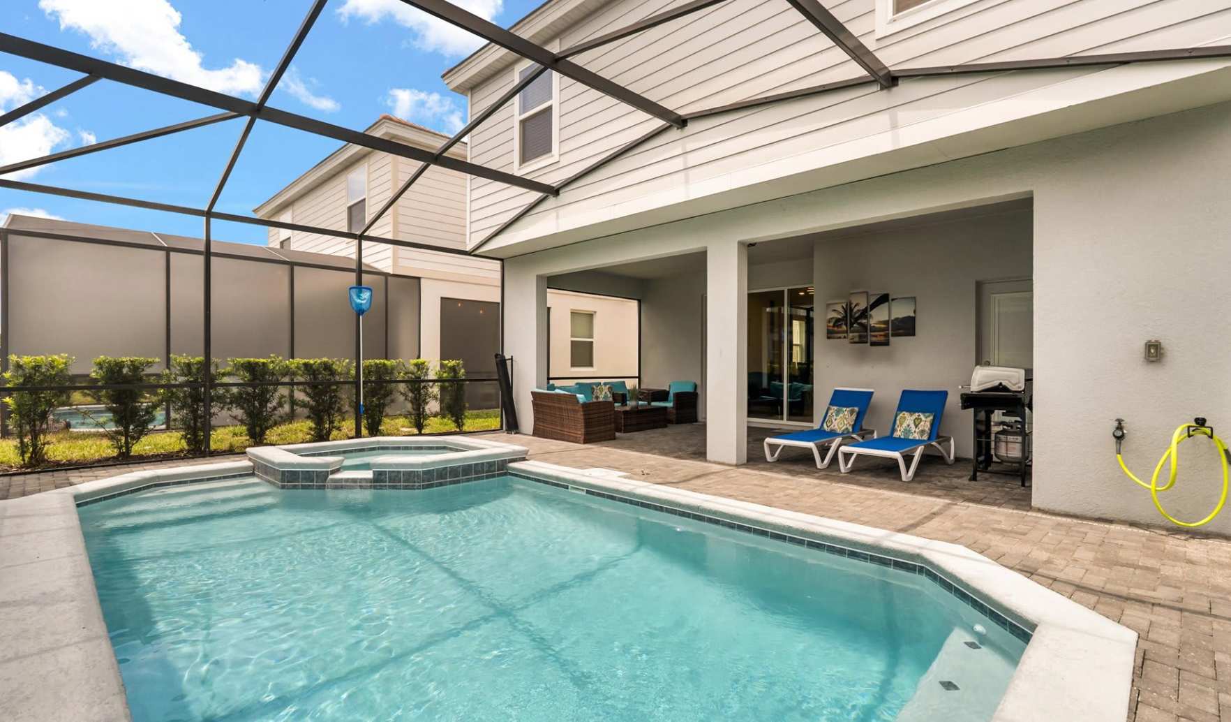 Expansive screened pool area featuring a rectangular swimming pool with an integrated hot tub, two blue lounge chairs, and a covered patio with a BBQ grill.