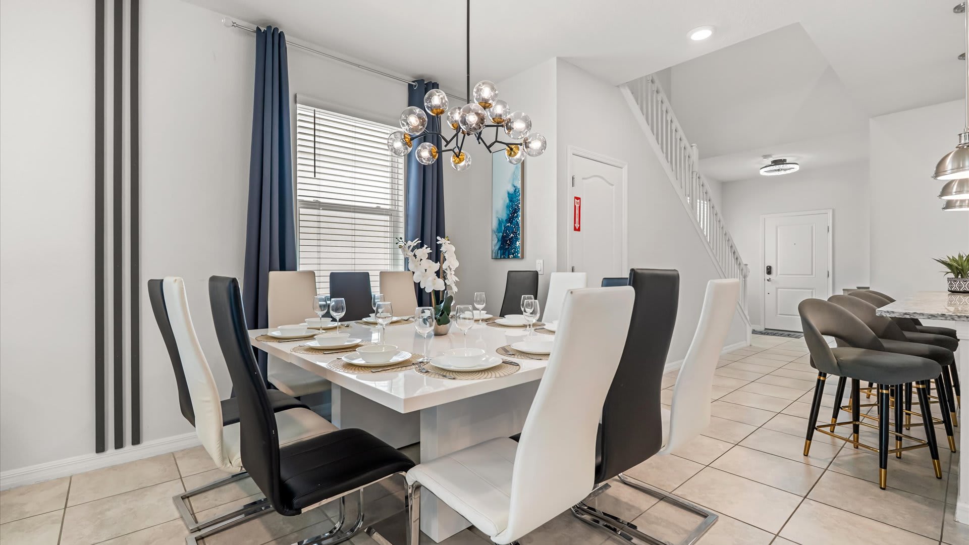 Spacious dining area with a white square table and black and white chairs, illuminated by a modern chandelier, with a view towards the kitchen island with bar stools.