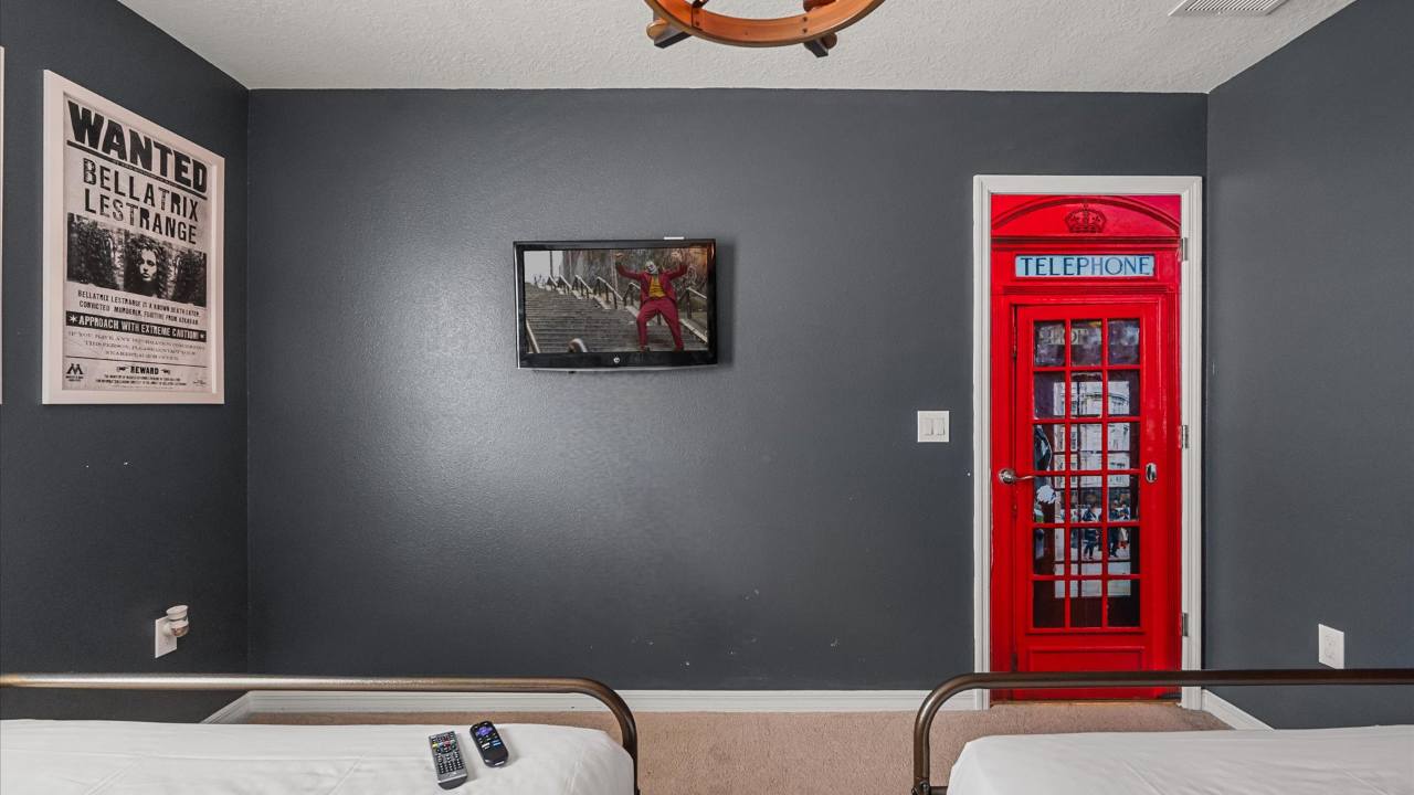 Themed bedroom with dark gray walls, featuring a "Wanted" poster, a wall-mounted TV, and a vibrant red telephone booth decal on a door, with two single beds partially visible in the foreground.