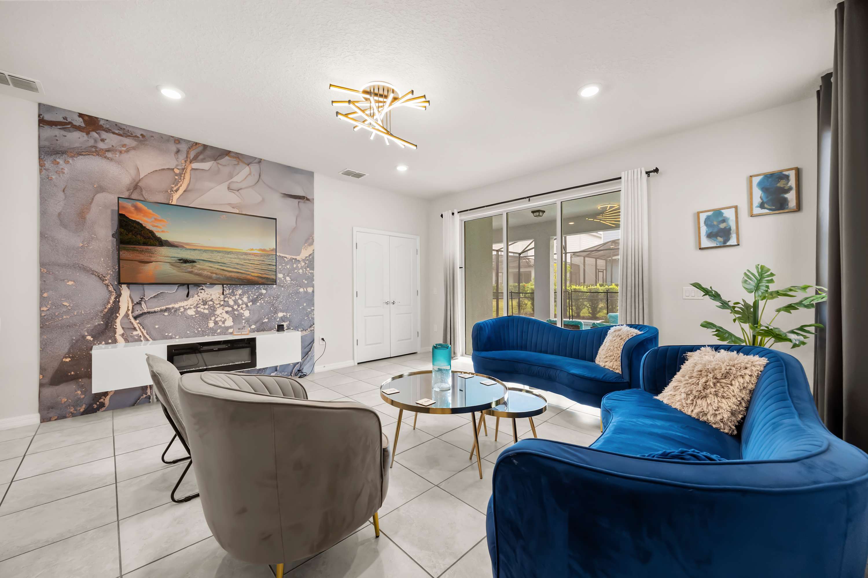 Bright living room with a large TV on a marble-patterned accent wall, blue velvet sofas, and sliding glass doors leading to an outdoor space, all under recessed lighting.