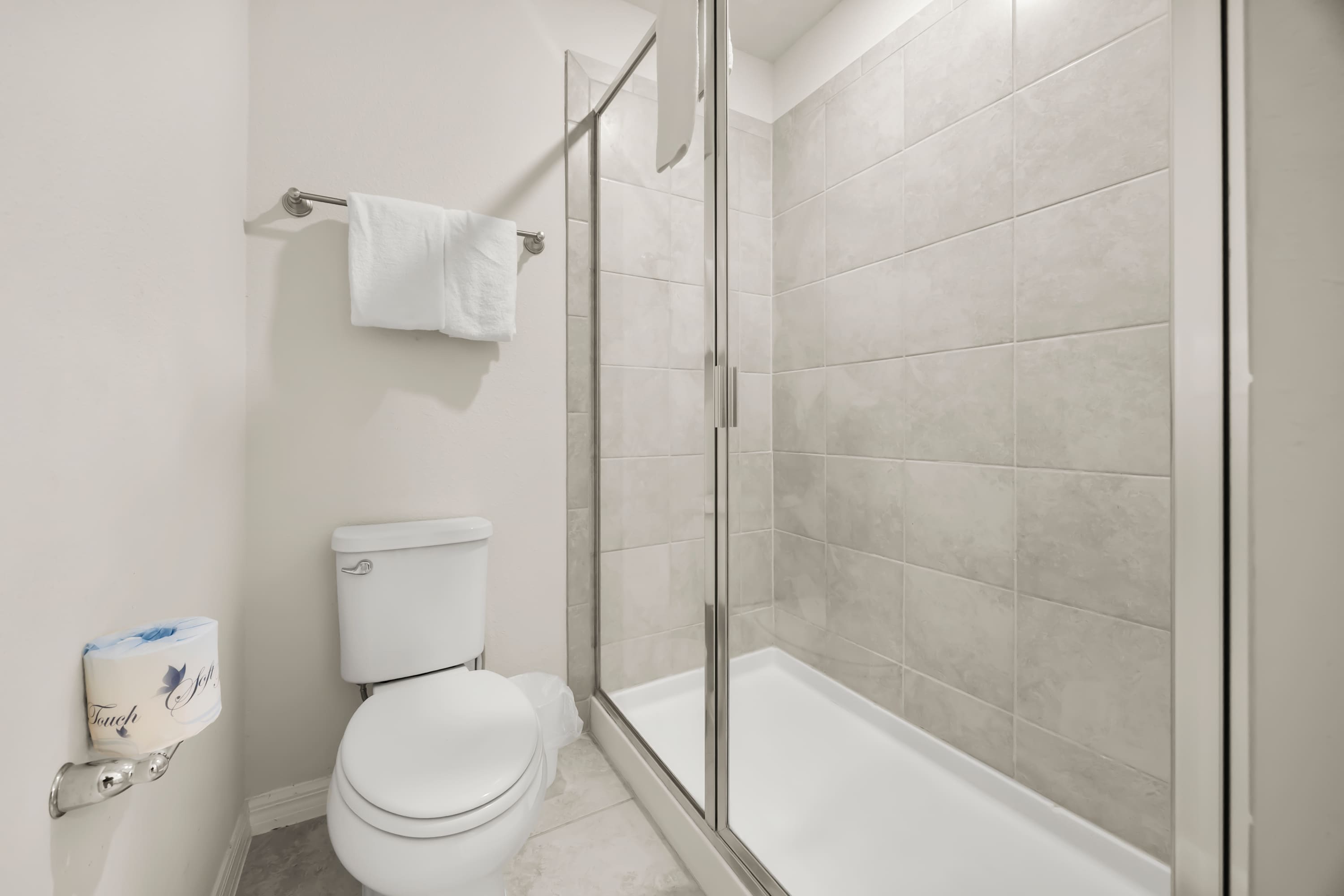 A compact modern bathroom featuring a white toilet, a glass-enclosed walk-in shower with light gray rectangular tiles, and a towel rack with white towels on a light-colored wall.