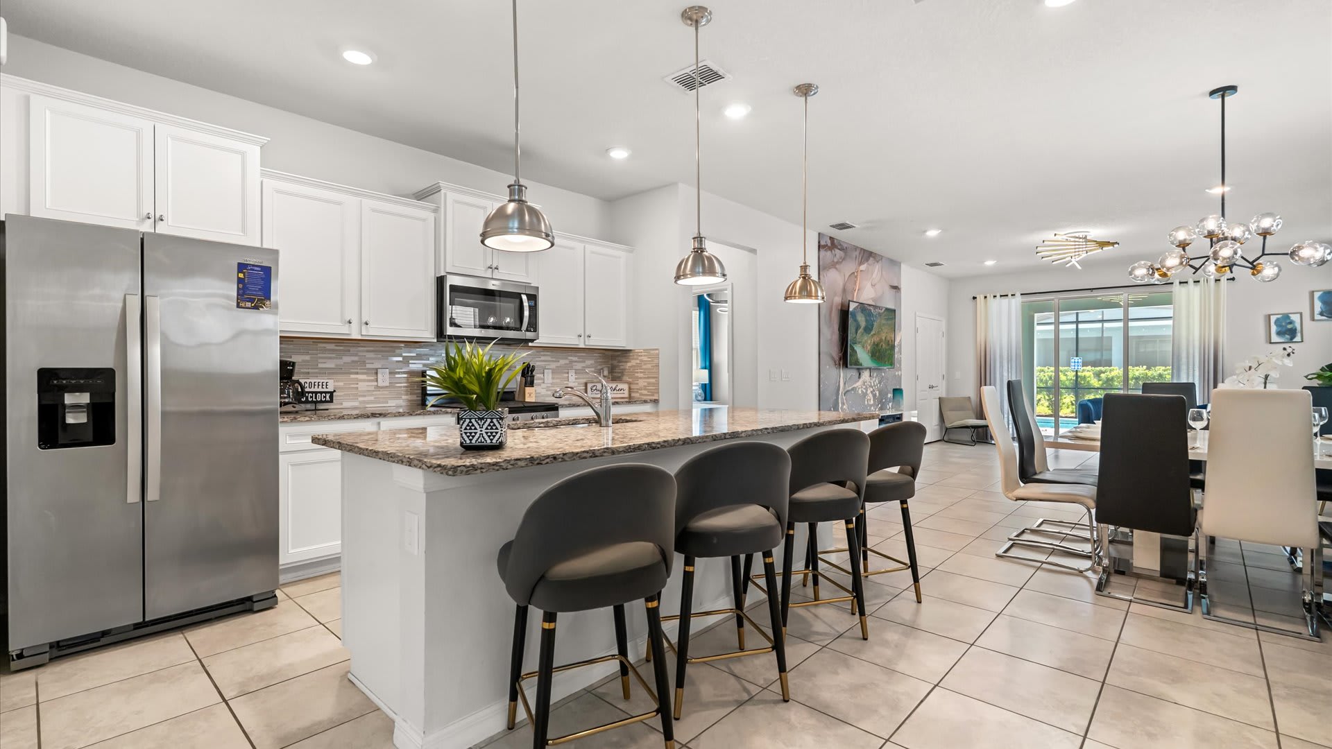 An open-concept kitchen with white cabinets, granite countertops, and a large island with bar seating, flowing into a dining area and living room with a view of the pool area.
