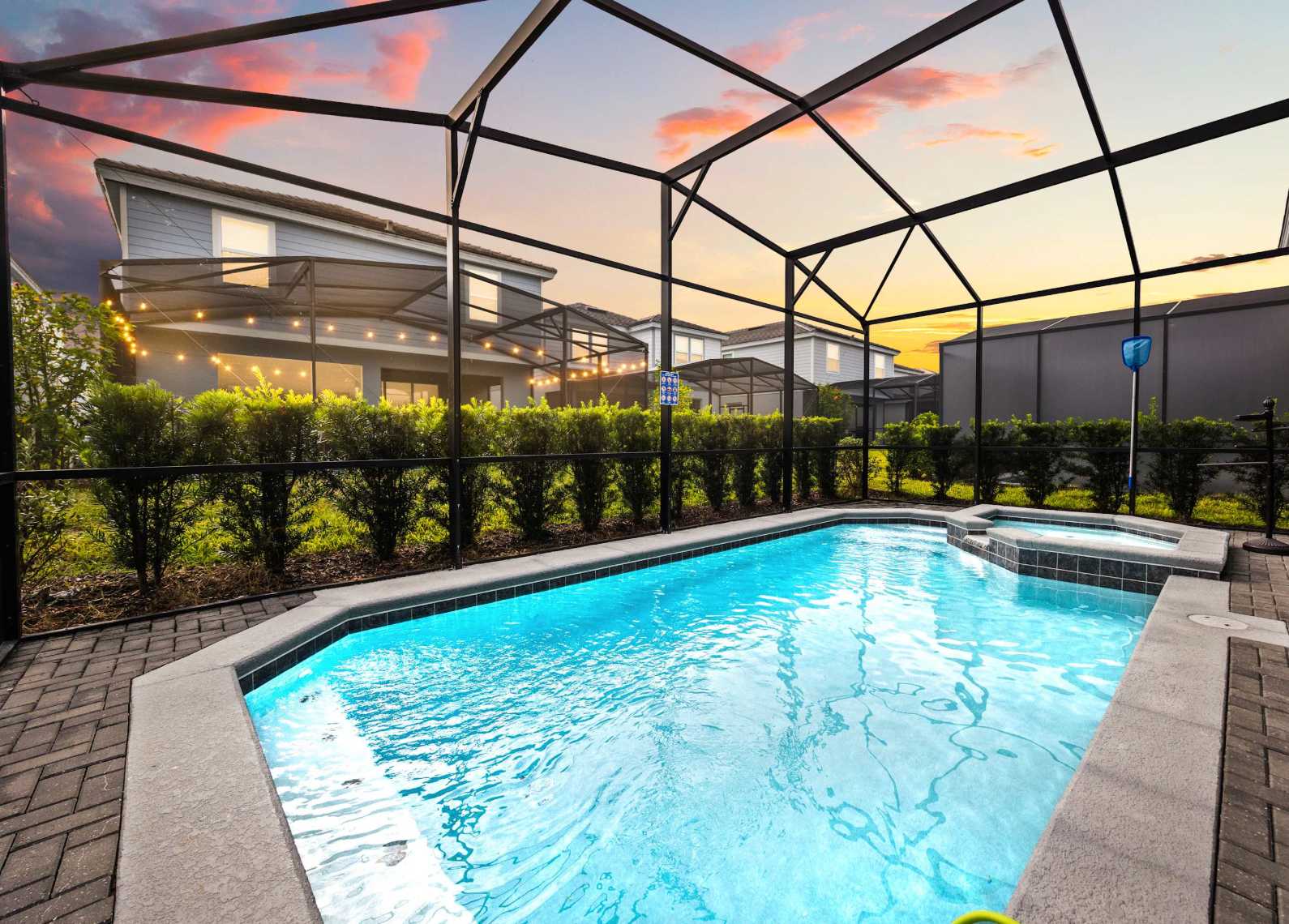 Private swimming pool and hot tub within a screened enclosure, illuminated by string lights, set against a vibrant sunset sky with neighboring houses visible in the background.