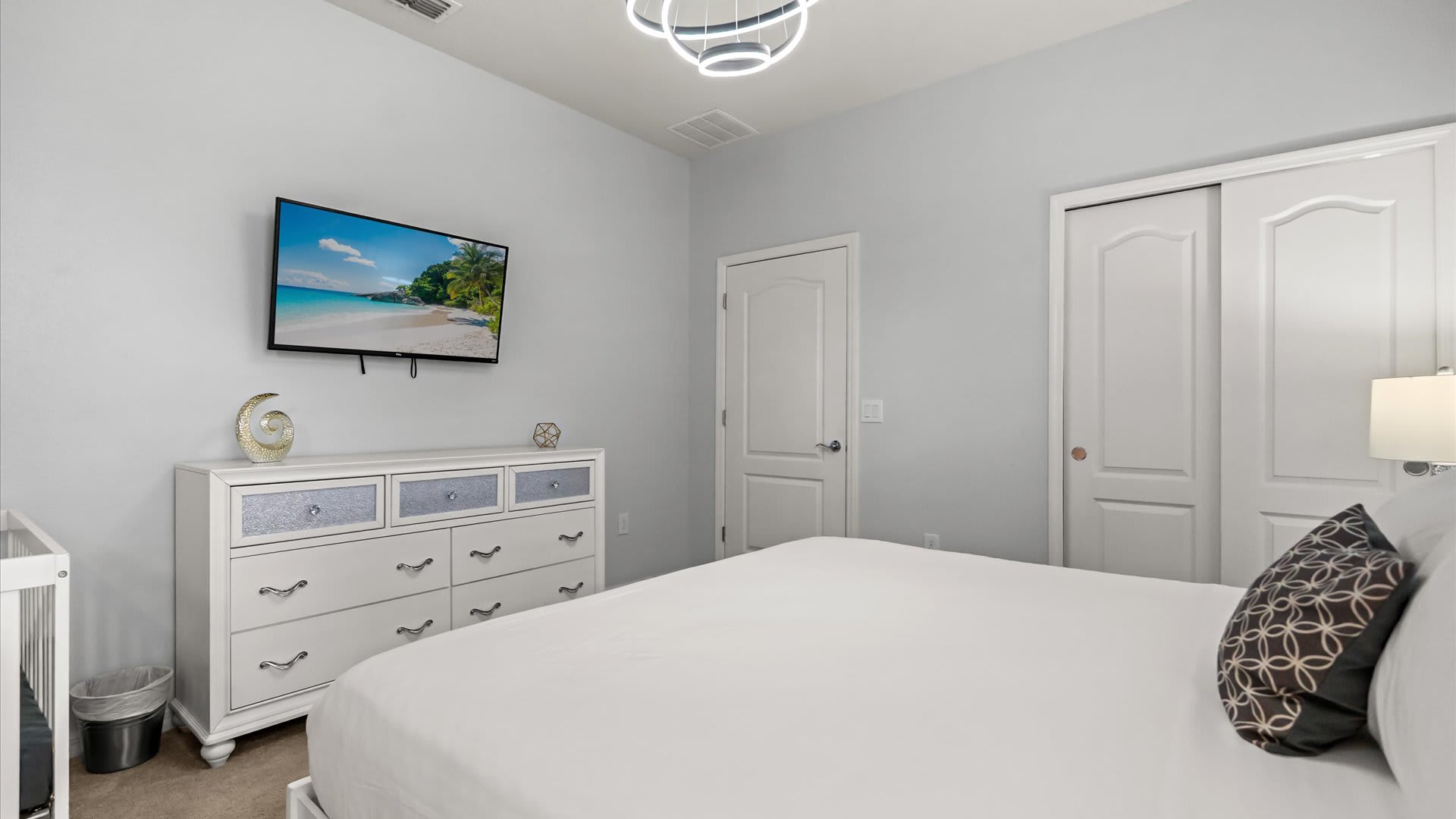 View of a bright bedroom showing a white dresser with a wall-mounted TV above it, a small crib, and a closet with bi-fold doors, maintaining a clean and modern aesthetic.