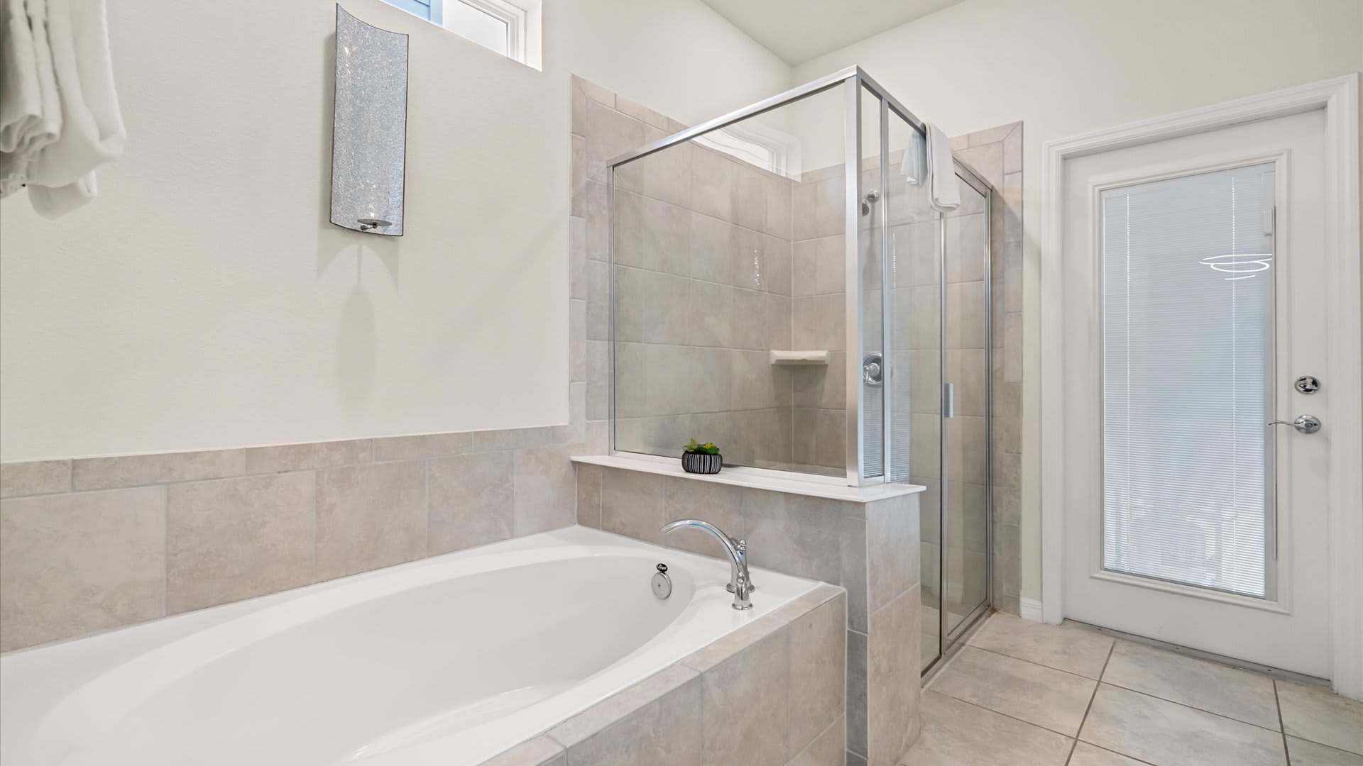 Spacious bathroom with a tiled built-in bathtub and a separate glass-enclosed walk-in shower, featuring light-colored tiles and a modern wall sconce.