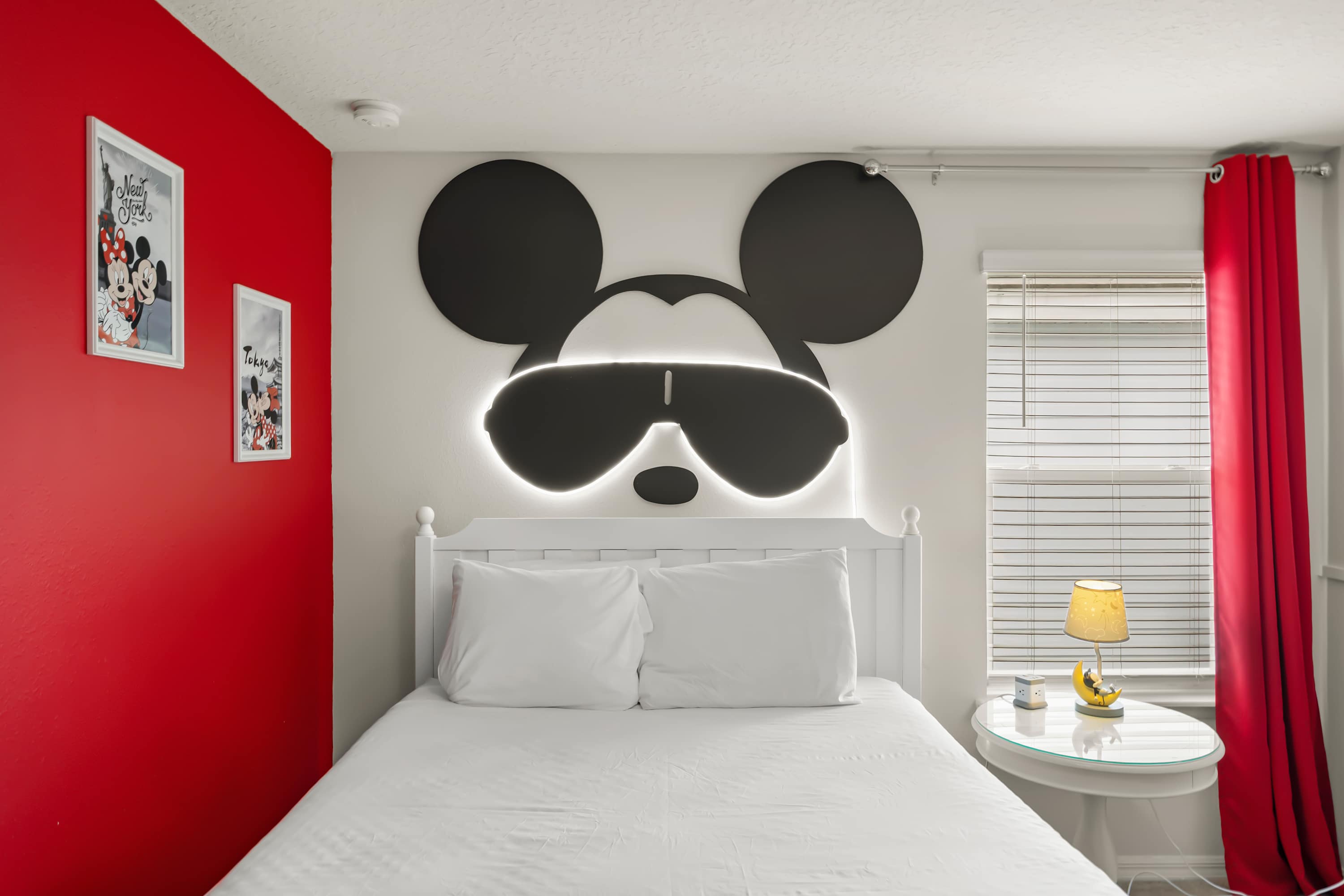 A bright Mickey Mouse themed bedroom featuring a white full-sized bed with a large illuminated Mickey headboard, a solid red accent wall, and a bedside table with a unique moon and character lamp.
