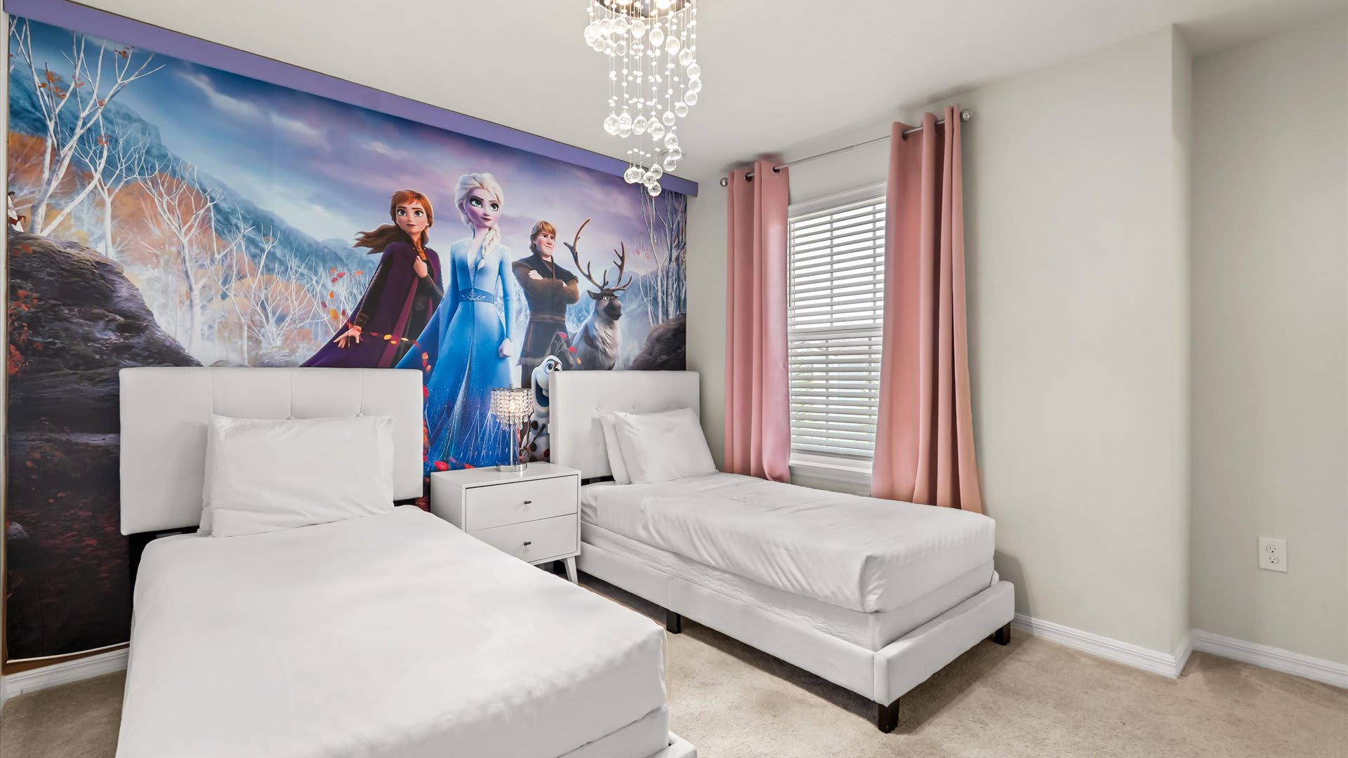 Frozen-themed bedroom with two white single beds and a shared nightstand, set against a large wall mural depicting Elsa, Anna, Kristoff, and Sven in a winter landscape, under a decorative chandelier.