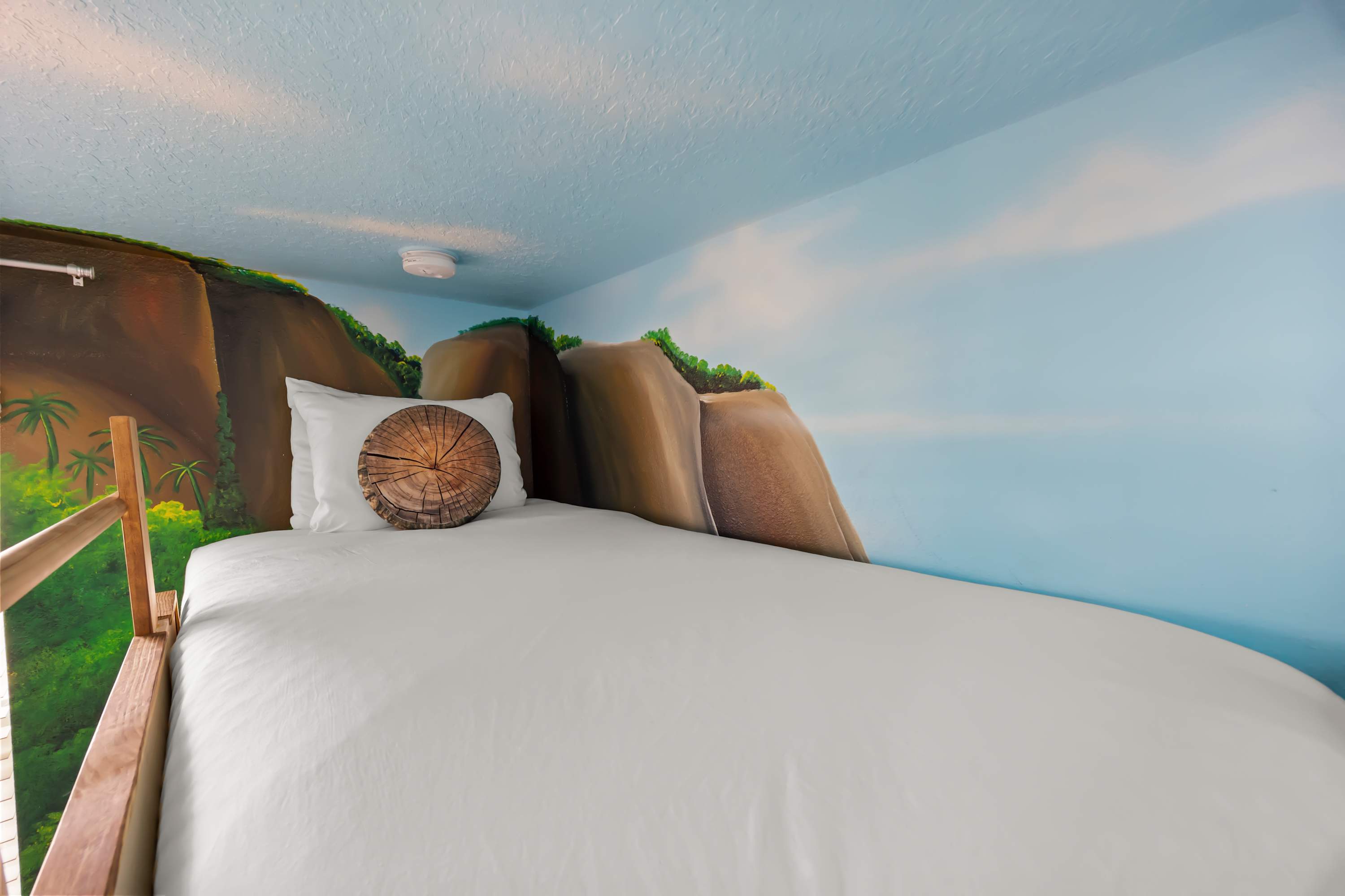 A close-up of the top bunk bed in the Moana-themed room, featuring a white mattress, a round wood-slice patterned pillow, and a painted mural of rocky cliffs on the wall.