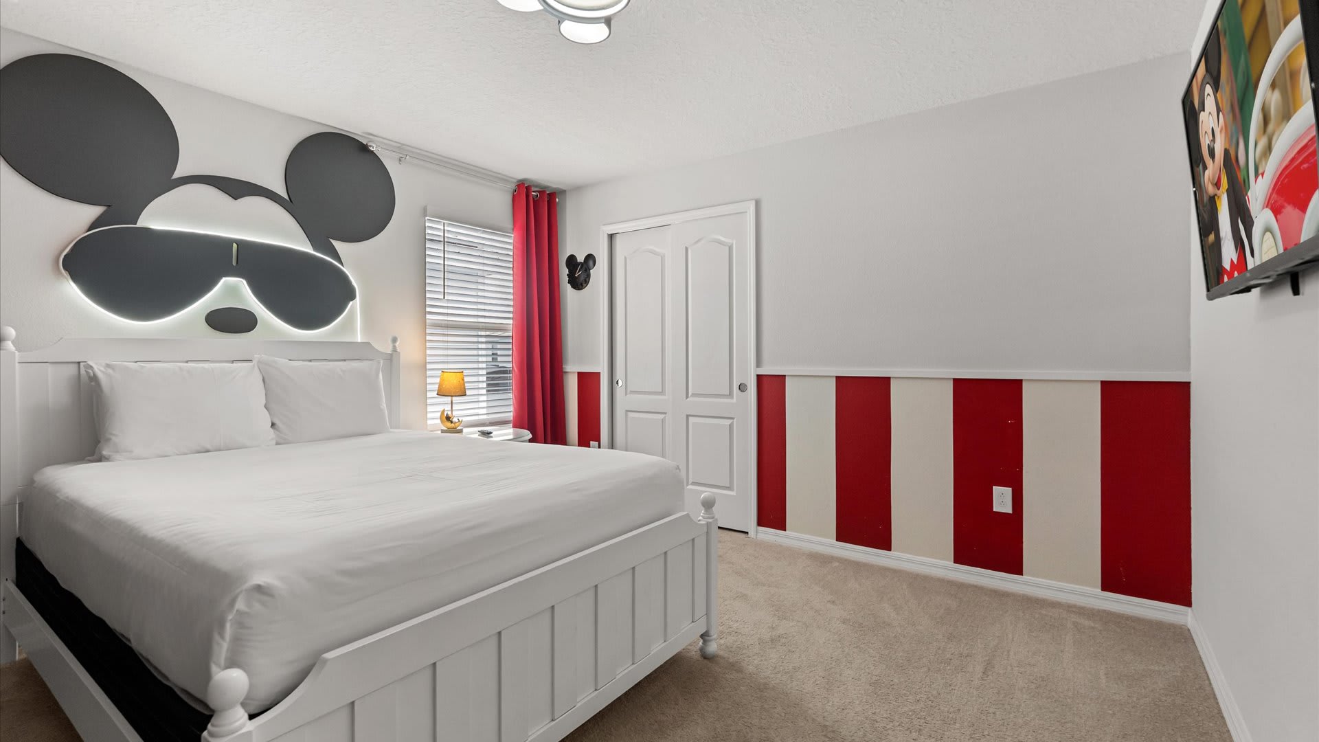 A vibrant Mickey Mouse themed bedroom featuring a white queen-sized bed with a large illuminated Mickey headboard, red and white striped wainscoting, and a wall-mounted TV displaying a cartoon.