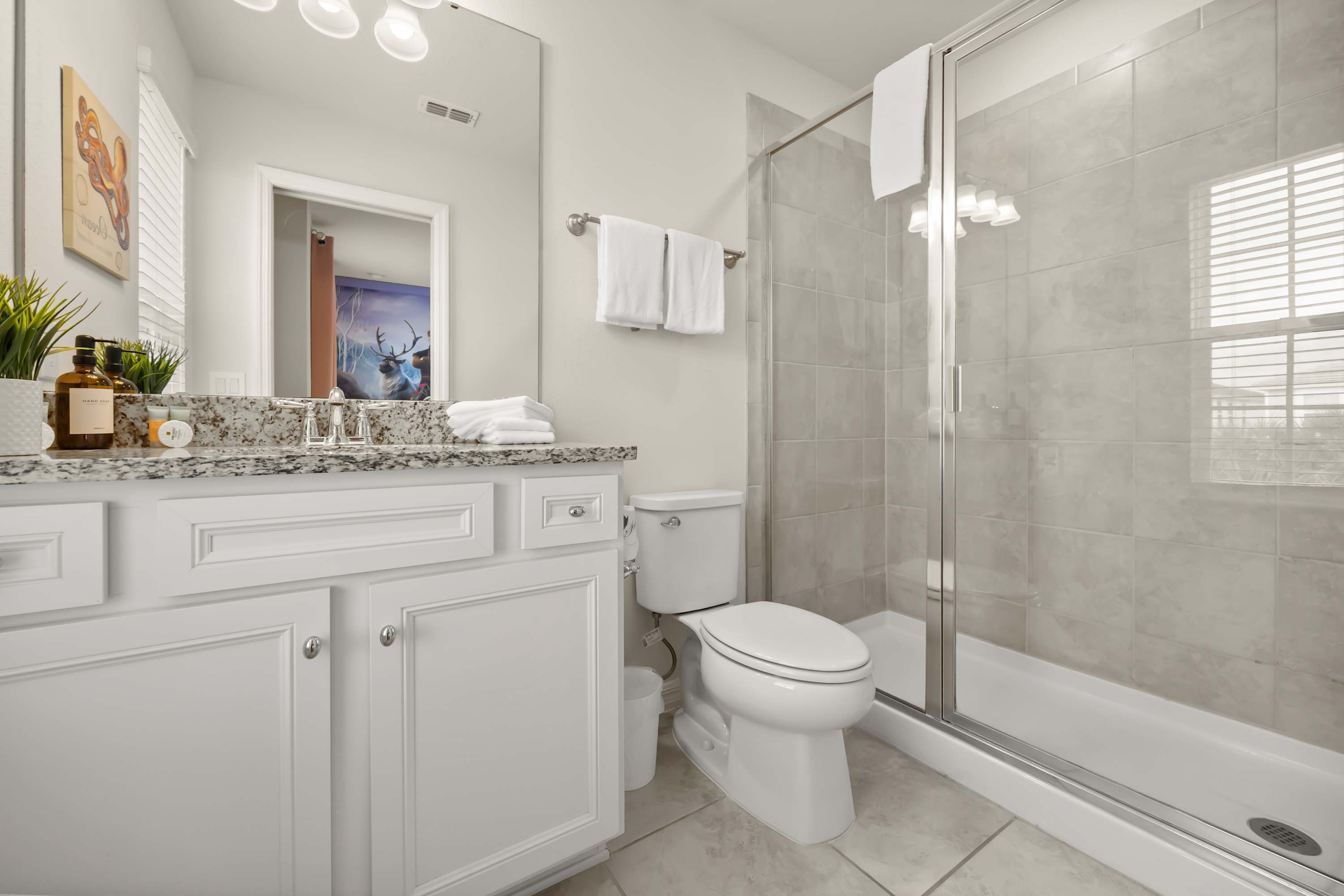 Bright bathroom with a white vanity featuring a granite countertop and a large mirror. A white toilet is next to a glass-enclosed walk-in shower with light gray tiles. White towels are neatly folded on the vanity and hanging on a rack.