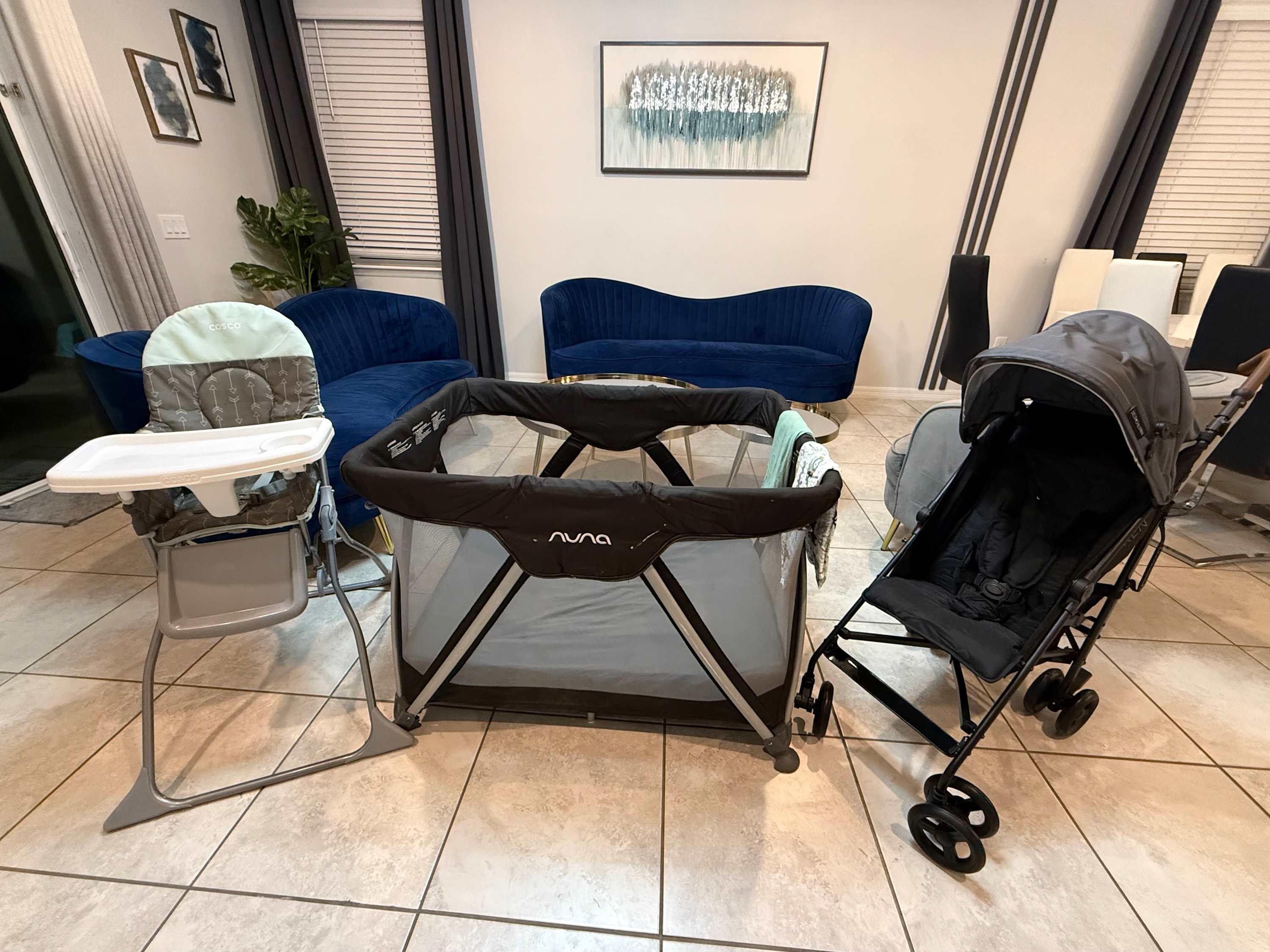 Bright living area featuring essential baby gear including a grey high chair, a black pack 'n play, and a grey stroller, set on light tiled flooring with blue velvet furniture in the background.