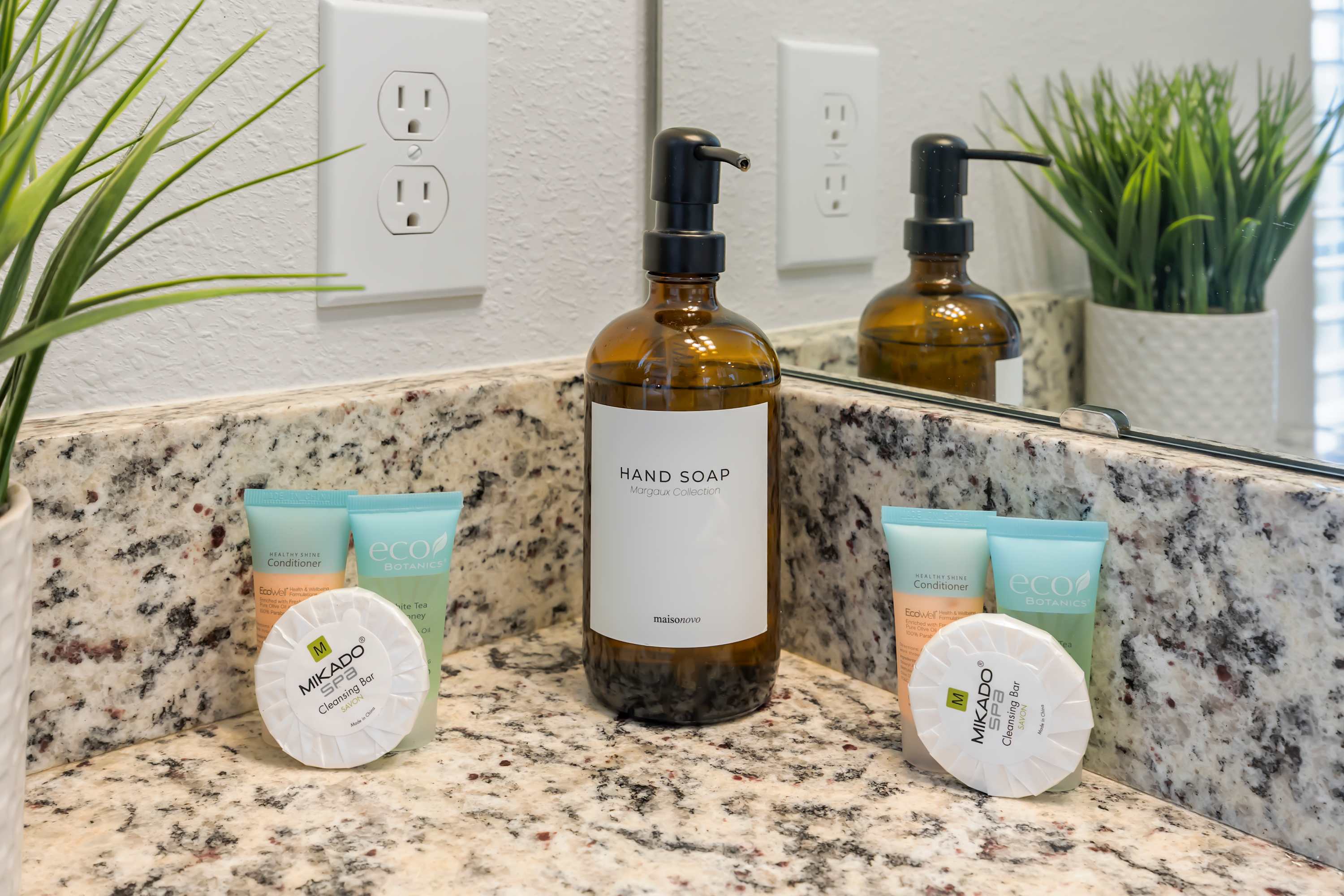 Close-up of a granite countertop with a large amber hand soap dispenser, two small bottles of conditioner, and two round bars of soap. A mirror reflects the items and a green potted plant, with two electrical outlets visible on the wall.