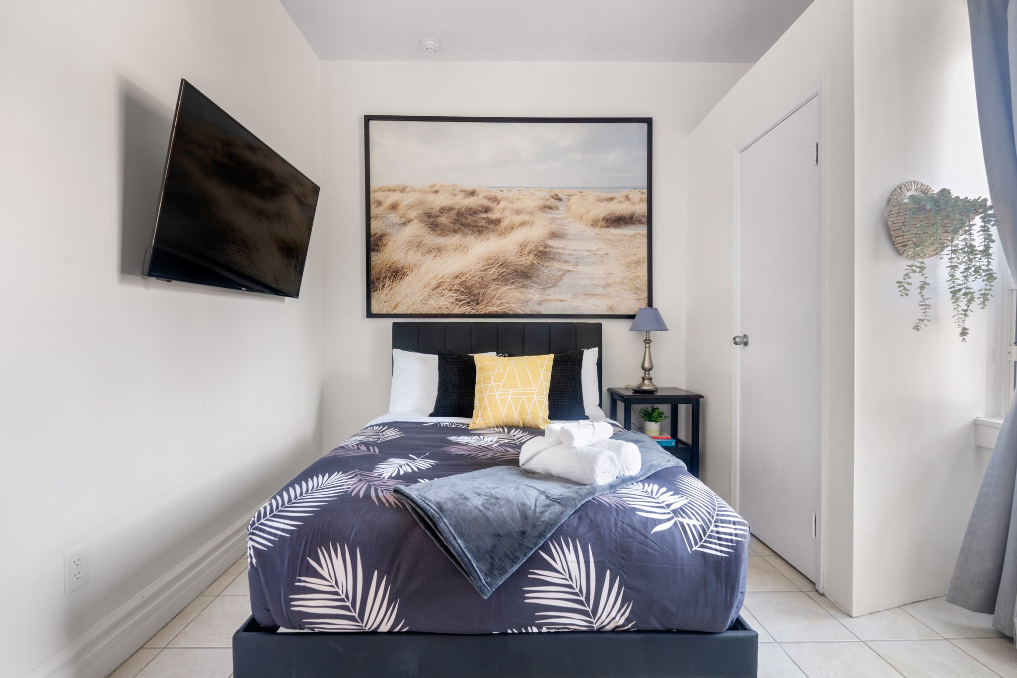 A close-up view of the black upholstered bed with a navy and white palm print duvet, white and yellow pillows, and neatly folded white towels. A large landscape art piece hangs above the bed, and a small nightstand with a lamp is to the right.