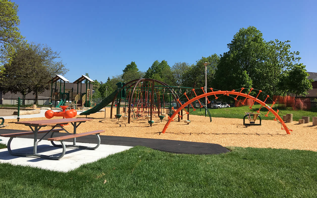 A vibrant outdoor playground under a clear blue sky, featuring various colorful climbing structures, slides, and swings on a wood chip surface. A picnic table is visible in the foreground, surrounded by green grass and mature trees.