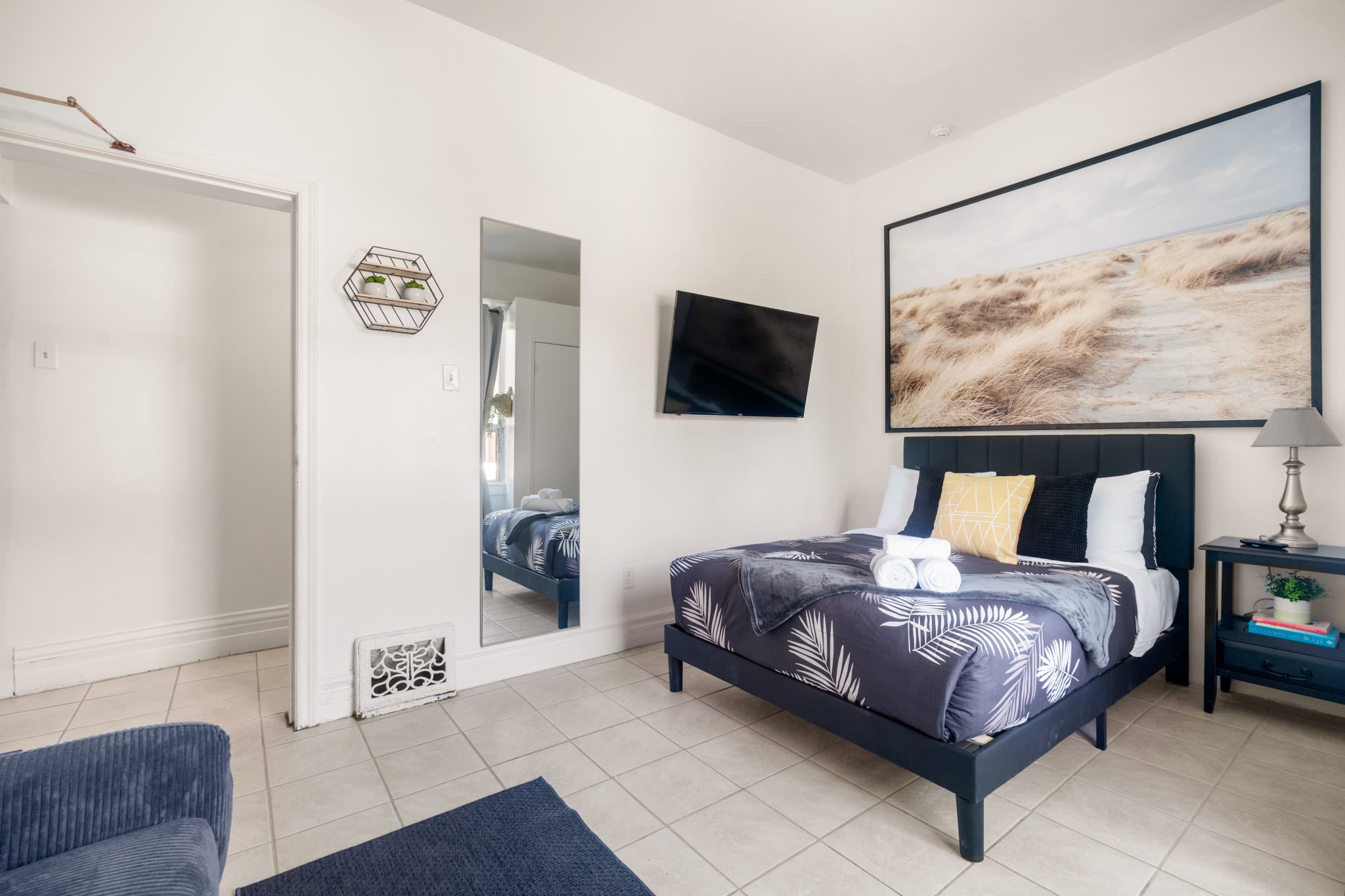 A cozy bedroom with a black upholstered bed, a navy and white palm print duvet, and a large landscape art piece above the headboard. A wall-mounted TV is visible to the left, and a full-length mirror reflects the room.