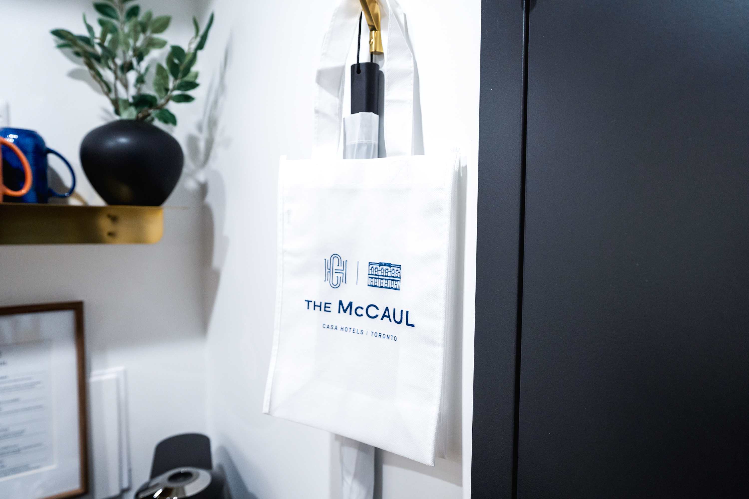 A white reusable tote bag with 'The McCaul CASA HOTELS | TORONTO' logo hanging on a hook, with a small plant and a coffee machine visible in the background.